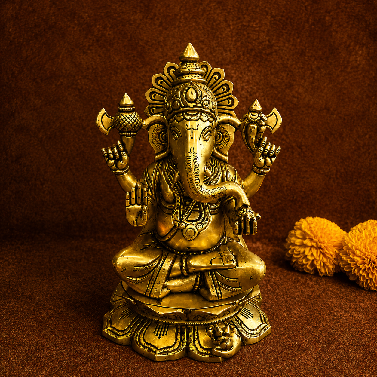 A brass Ganesha statue seated on a lotus base, photographed against a textured brown backdrop with two orange marigold flowers at the side.