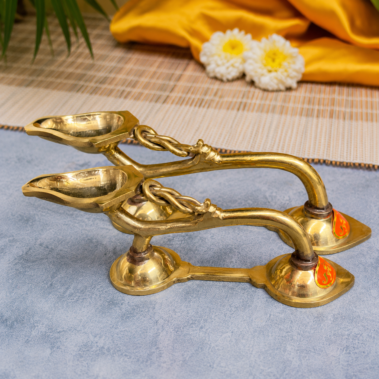 A pair of traditional brass oil lamps with decorative handles, displayed on a blue textured surface with a yellow fabric and white flowers in the background.