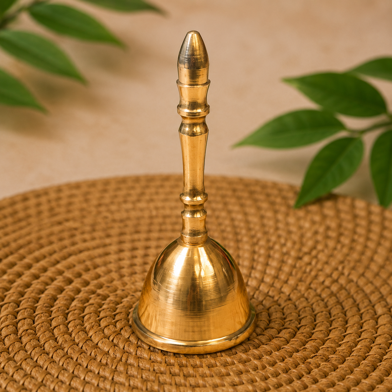 A polished brass bell with a decorative handle, standing upright on a round woven straw placemat with green leaves in the background.