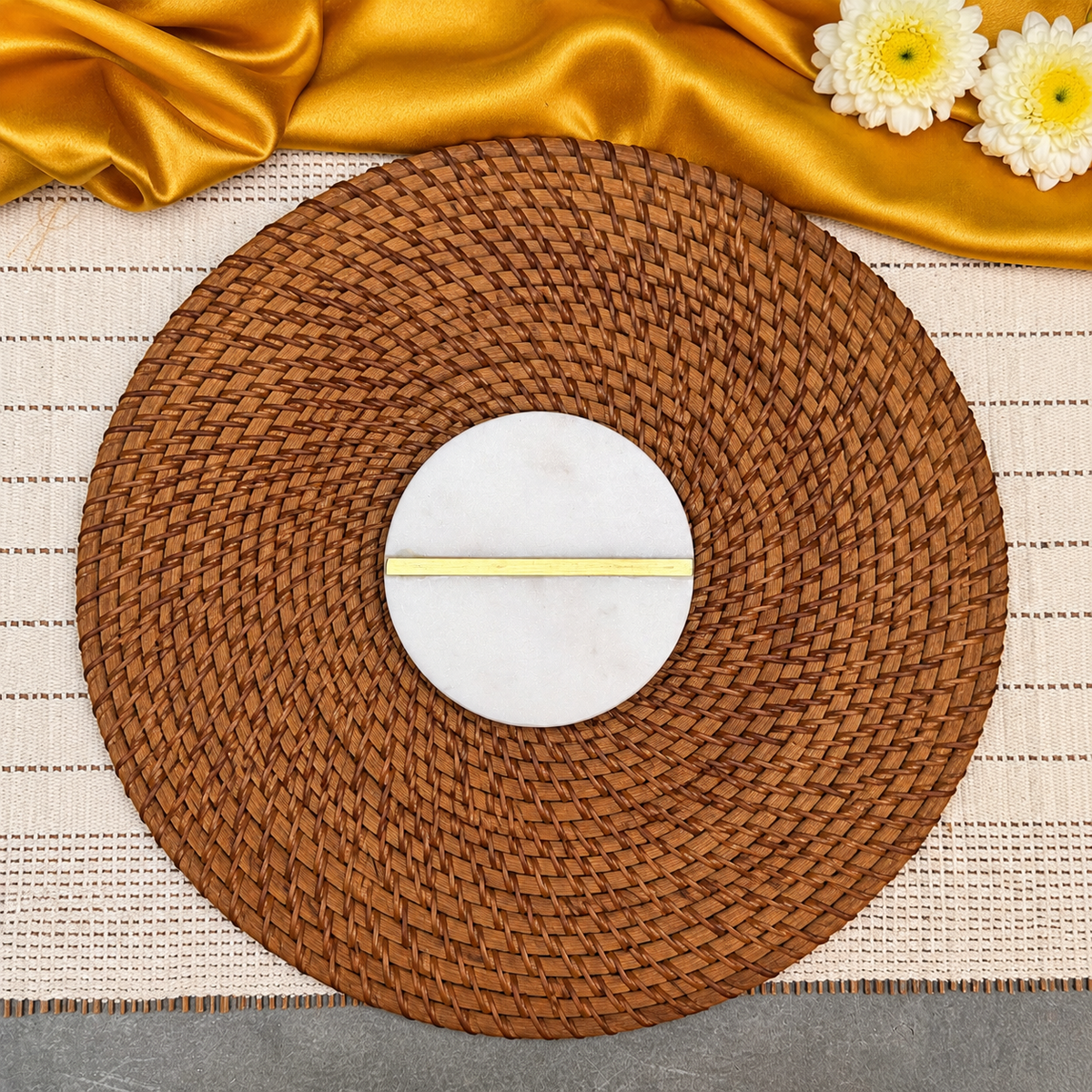 A round brown woven placemat with a white circular center and a thin gold horizontal stripe, styled on a beige woven runner with gold satin fabric and white flowers at the top.