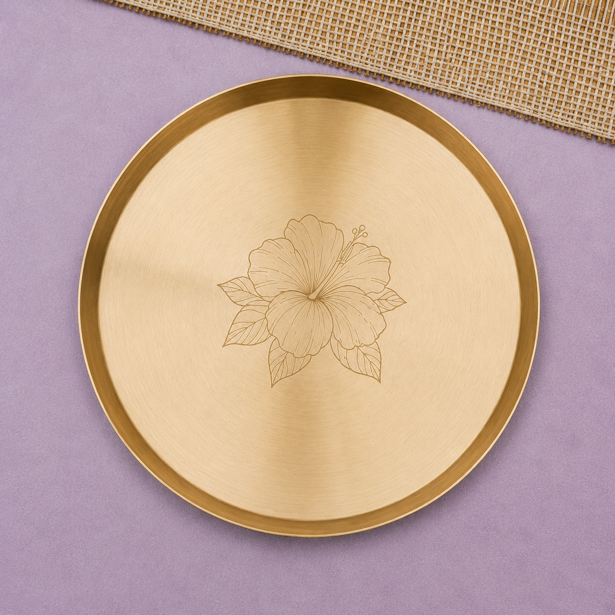 A round brushed brass tray with a polished gold rim and an engraved hibiscus flower in the center, displayed on a lavender background with a burlap ribbon in the corner.