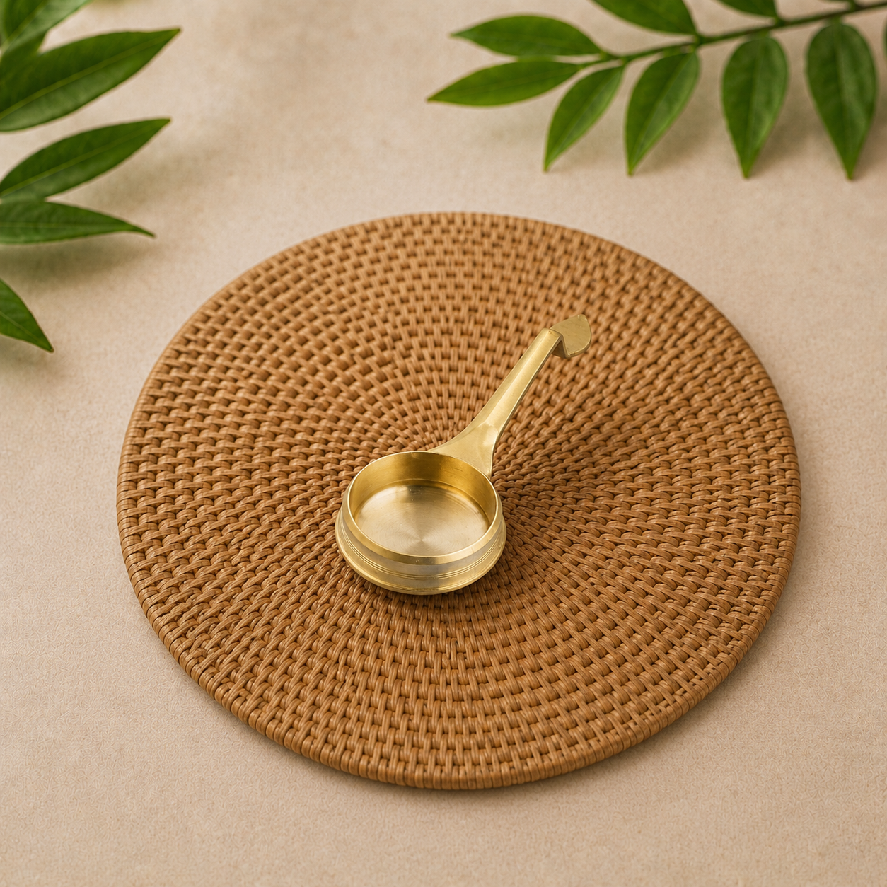 A round woven brown placemat on a beige surface, with a small gold-toned metal spoon resting on top and green leaves in the corners.