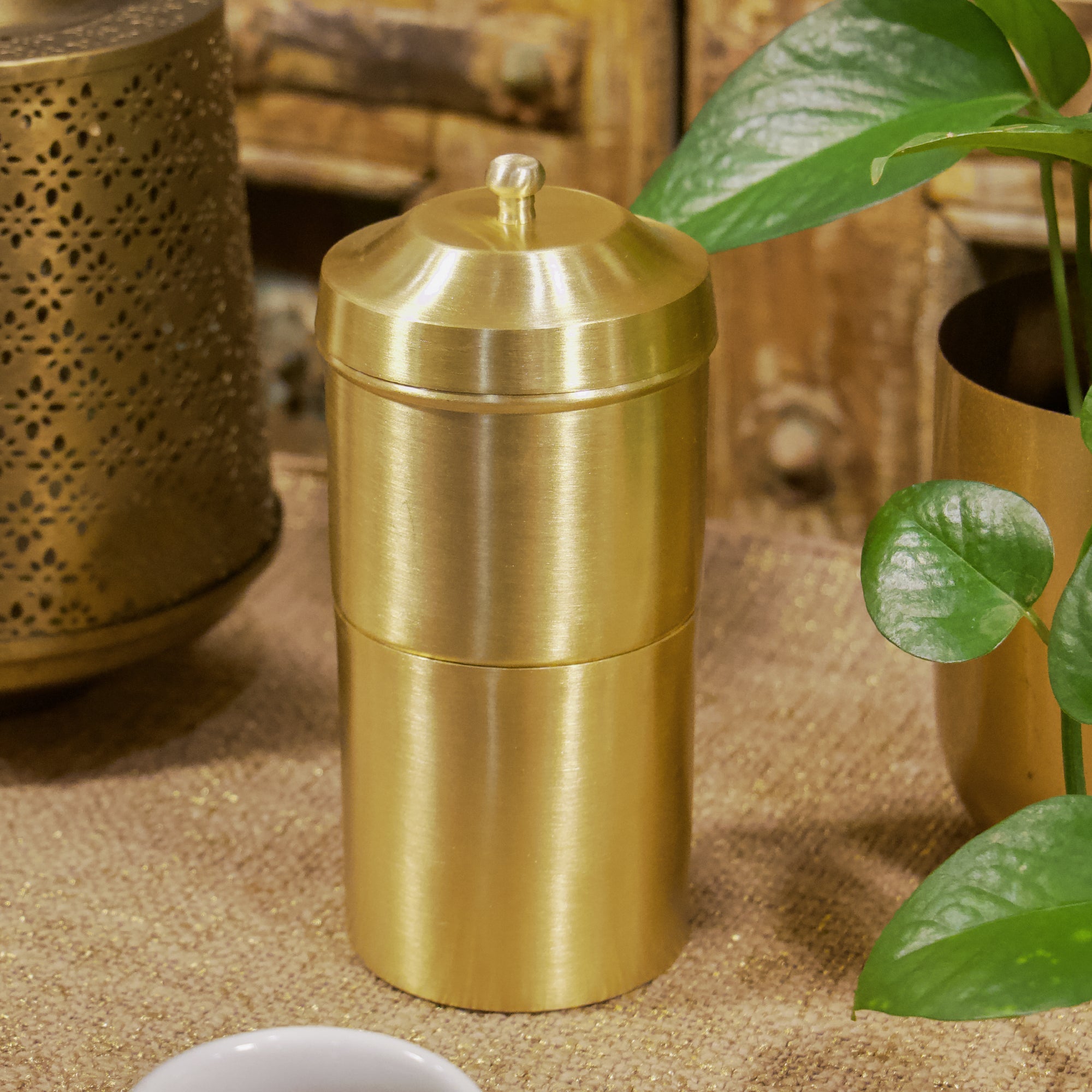 Gold cylindrical container on a textured surface with a plant in the background