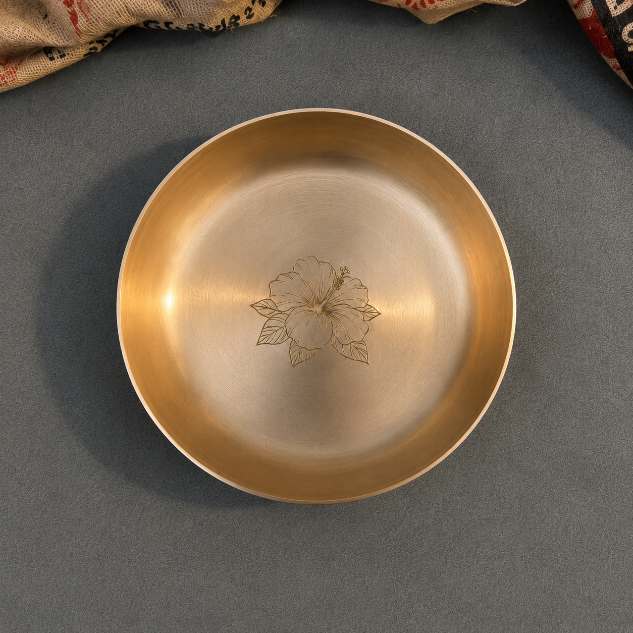 A round brushed metal dish with a warm gold-toned rim and a central floral engraving, photographed from above on a dark gray textured surface.