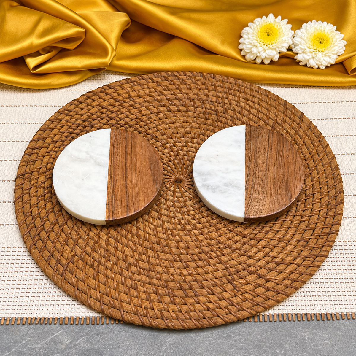 Two round coasters with a split white marble and dark wood design, arranged on a round woven brown placemat with gold fabric and white flowers in the background.