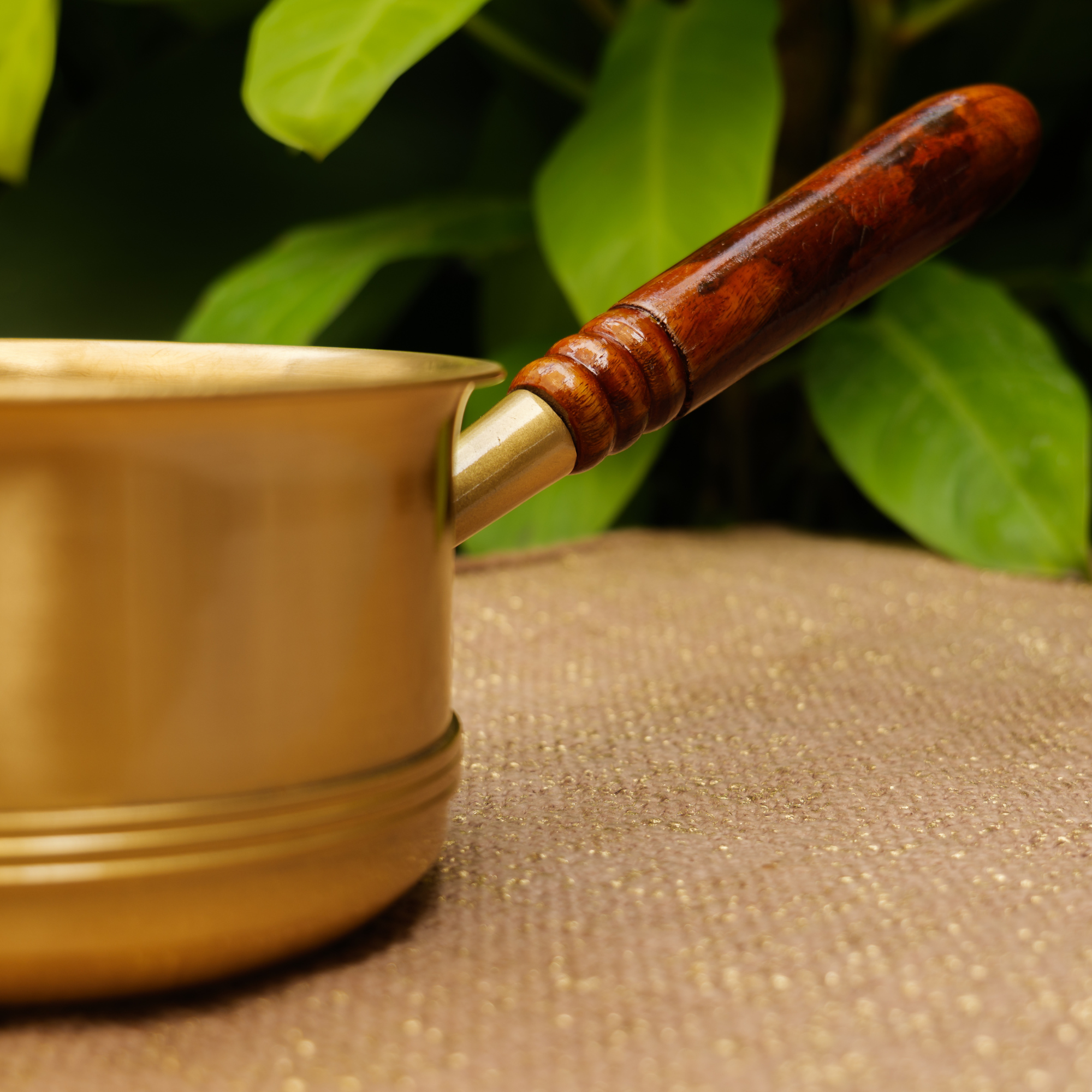 Brass pot with wooden handle on a textured surface with green leaves in the background