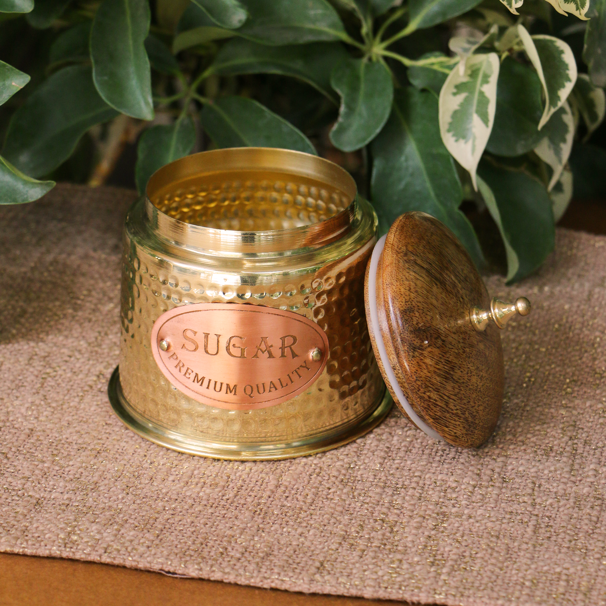 Vintage-style brass sugar container with wooden lid on a textured surface with green leaves in the background
