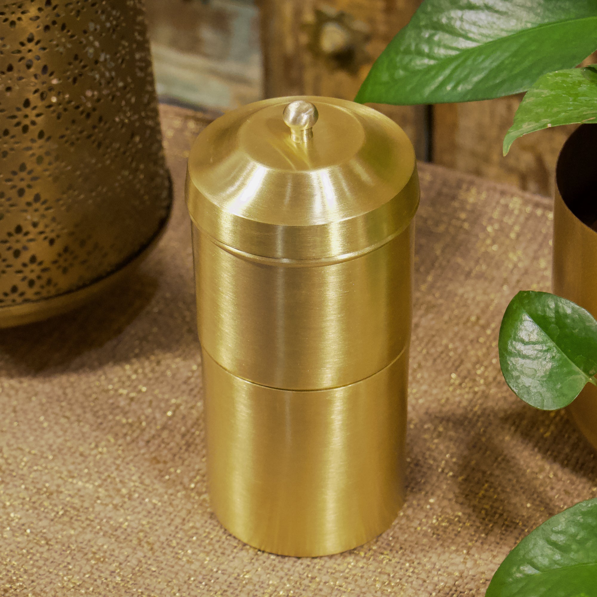 Gold metal container on a textured surface with plants in the background