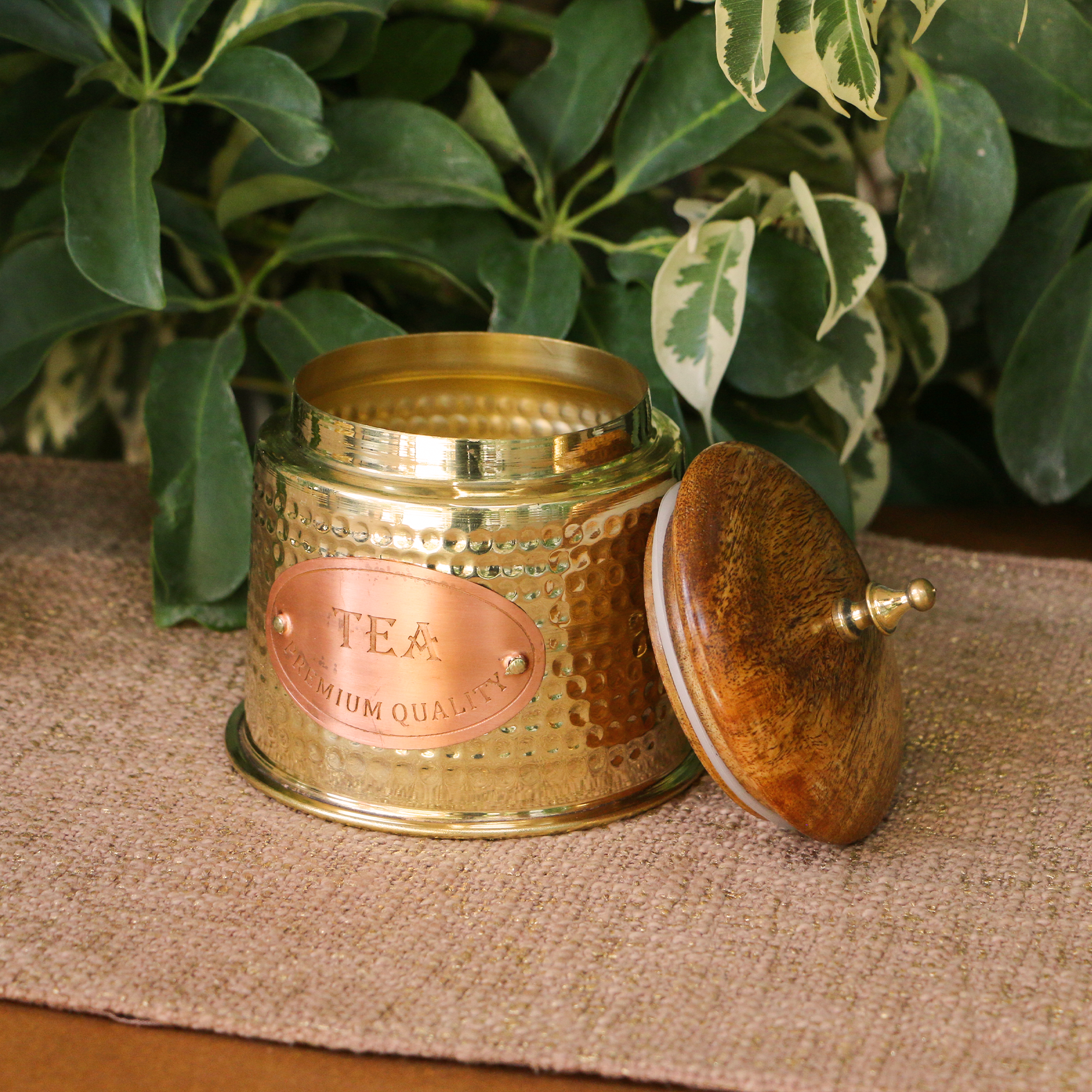 Gold tea canister with wooden lid on a textured surface with green leaves in the background