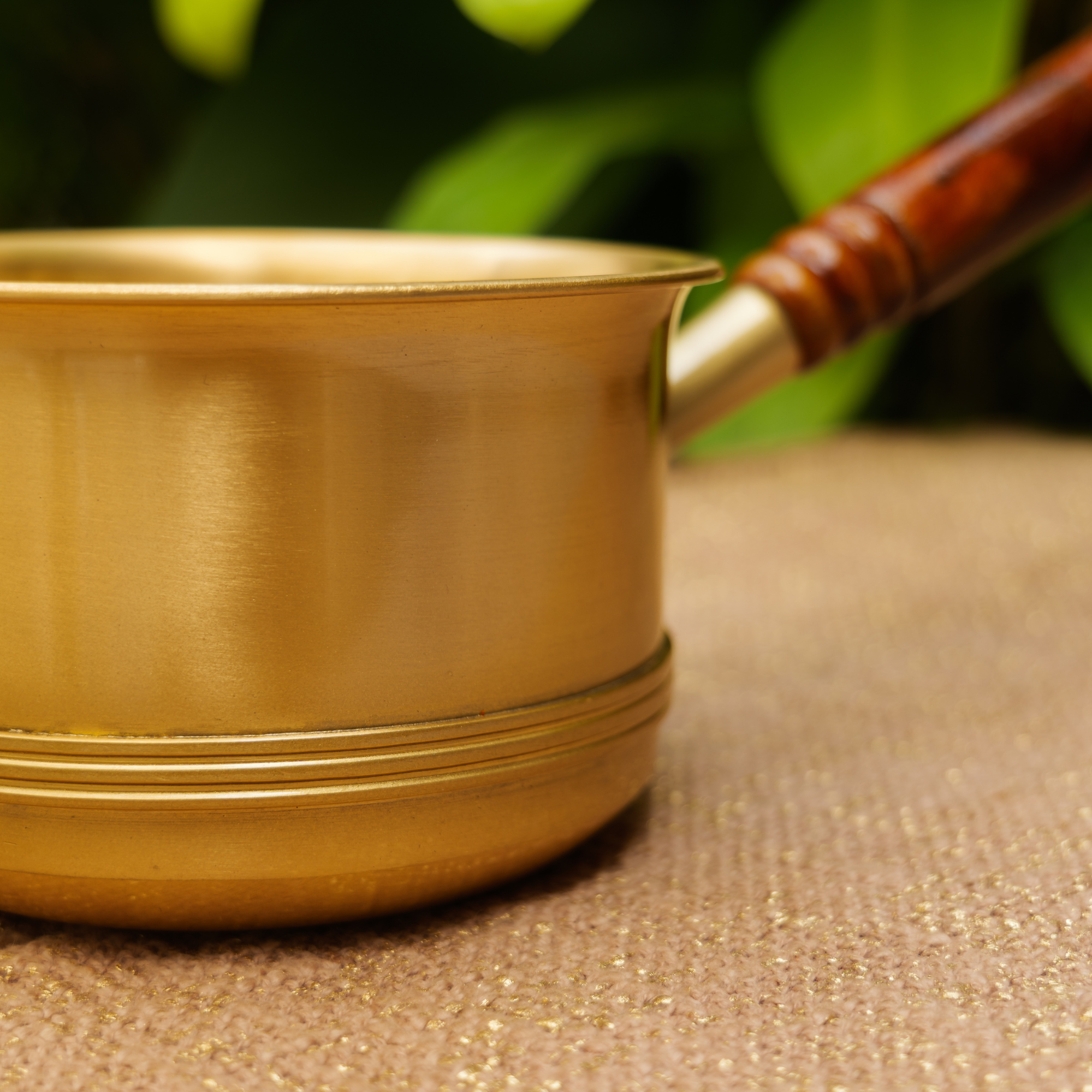 Brass pot with a wooden handle on a textured surface with green foliage in the background