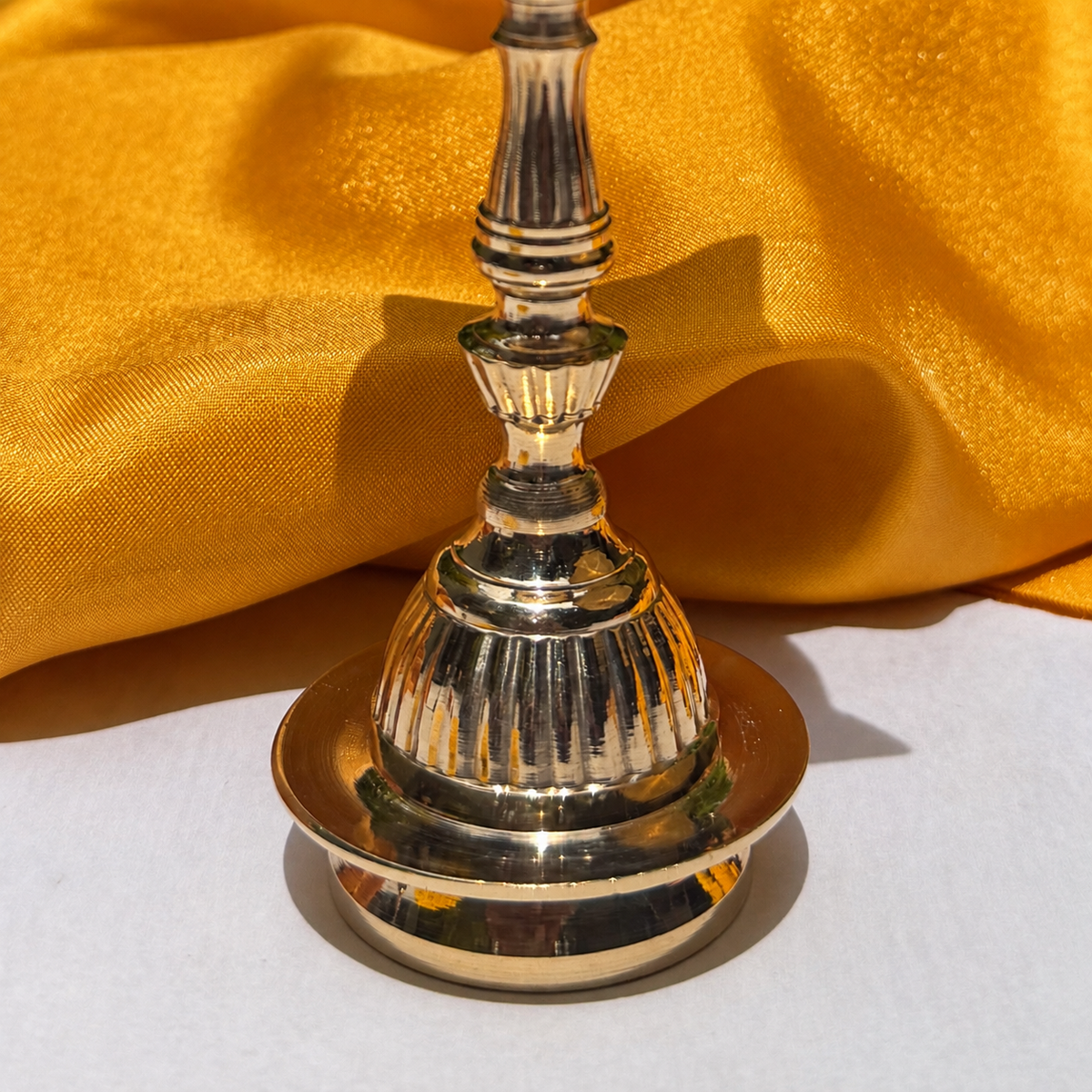 A polished brass or gold-toned metal bell with a fluted stem and circular base, photographed against draped golden fabric and a white surface.