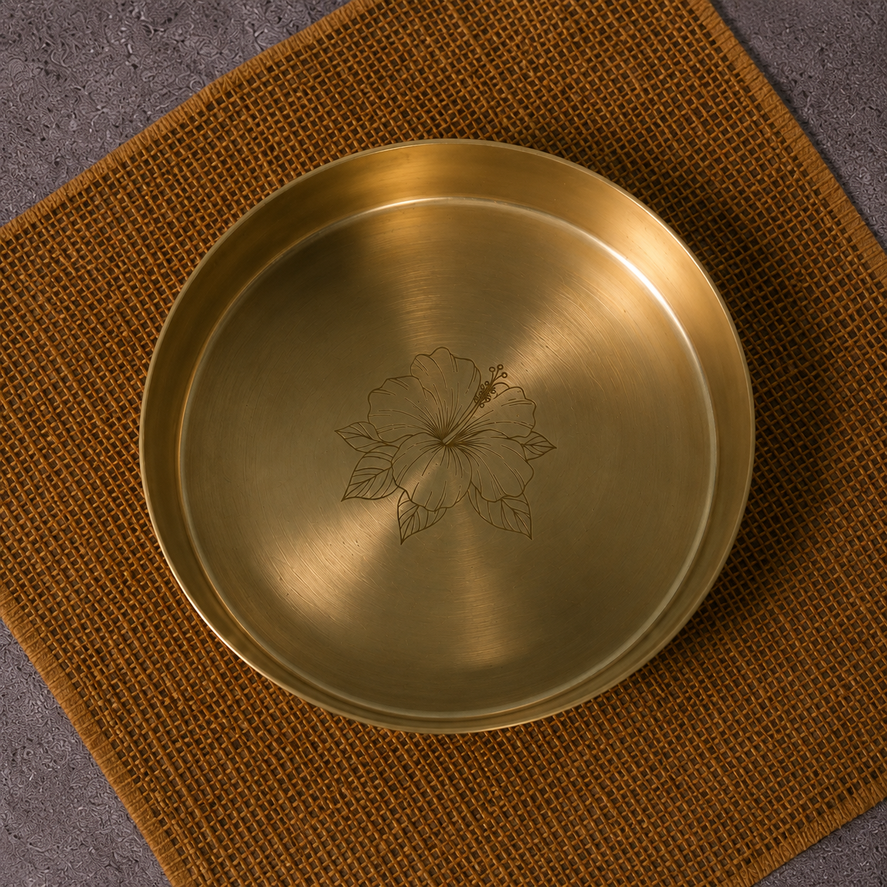 A round brushed brass plate with a floral engraving in the center, resting on a textured brown woven placemat over a gray surface.