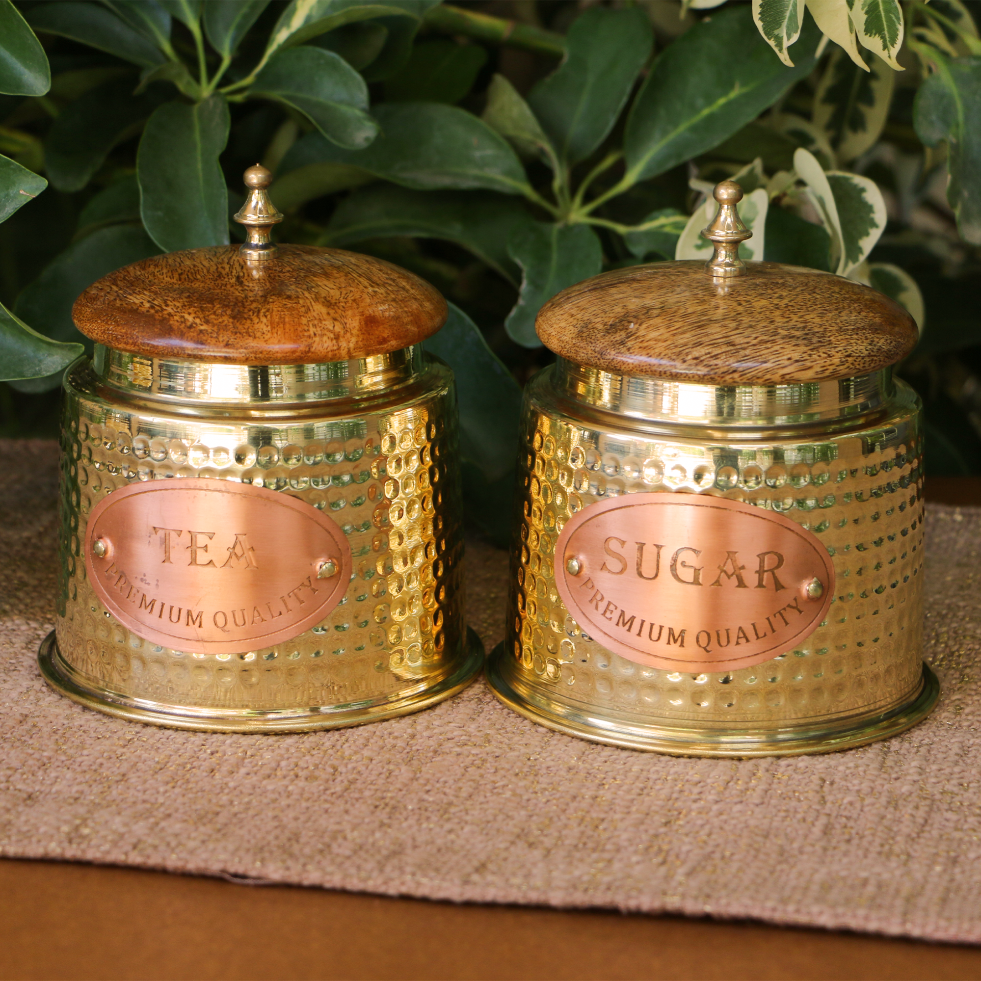Two gold tea and sugar canisters with wooden lids on a textured surface with green leaves in the background.