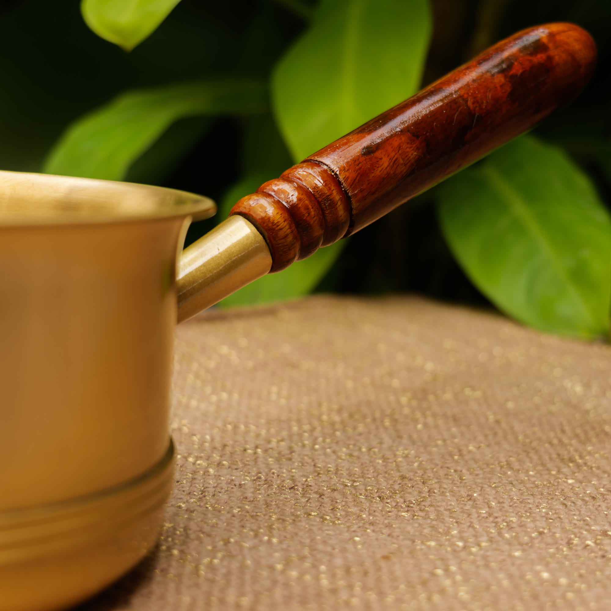 Brass pot with wooden handle on a textured surface with green leaves in the background