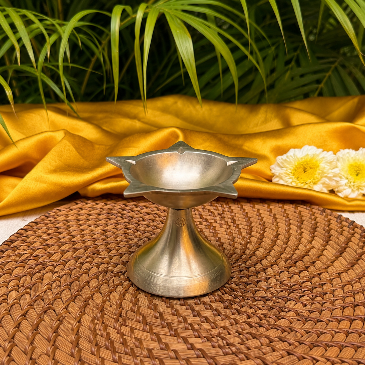 A silver-toned metal diya holder with a lotus-shaped top, displayed on a brown woven placemat with draped golden fabric, green leaves, and white flowers in the background.