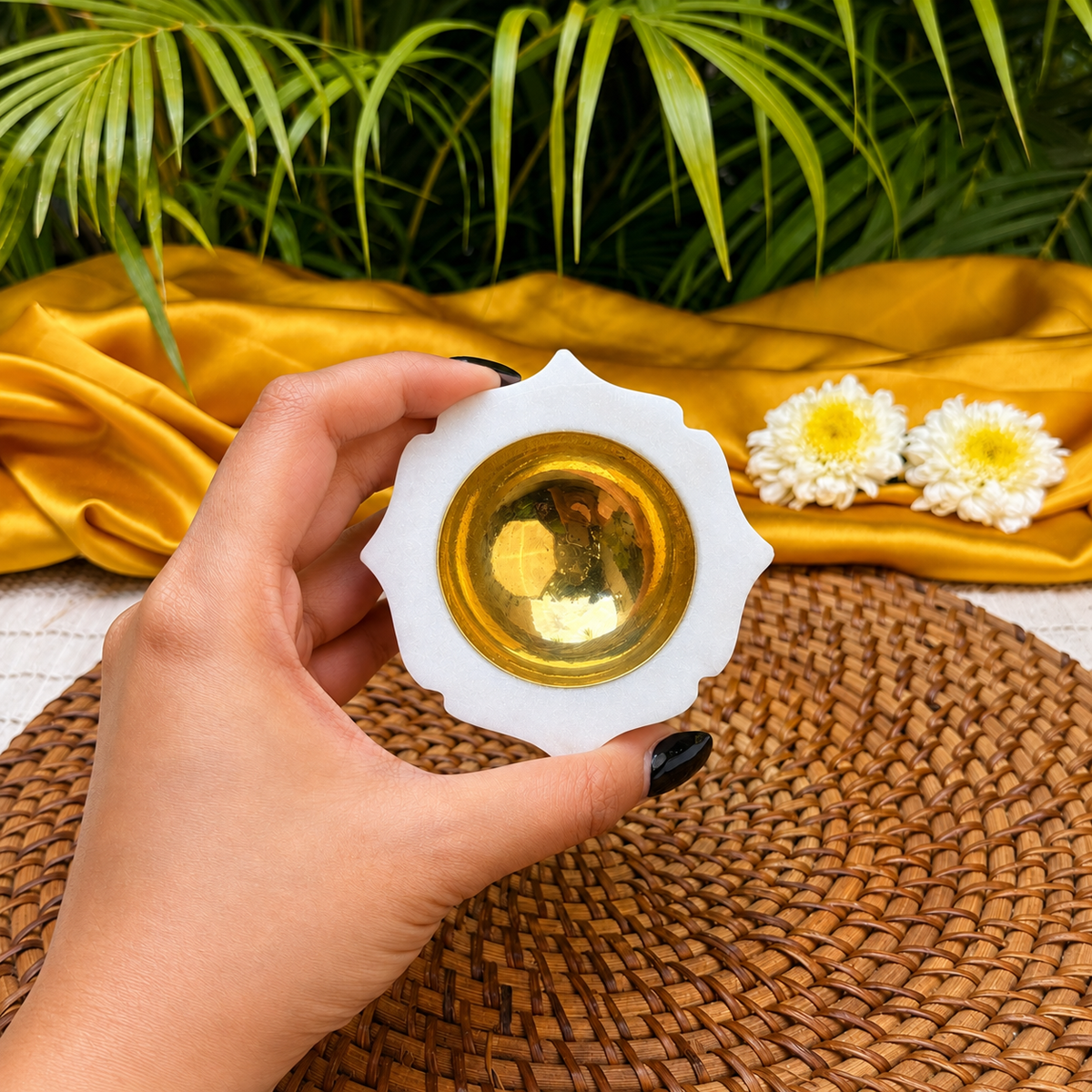 A hand with black nail polish holds a white lotus-shaped candle holder with a gold-toned glass center, set against a background of yellow fabric, white flowers, and green palm leaves.