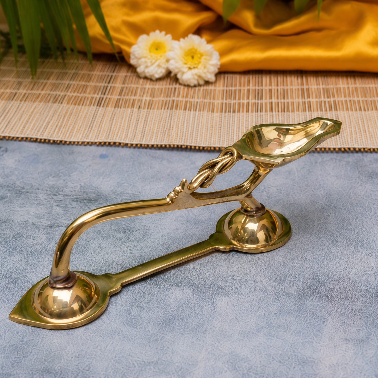 A polished brass oil lamp holder with two lamps, displayed on a textured blue surface with a bamboo mat, yellow fabric, and white flowers in the background.