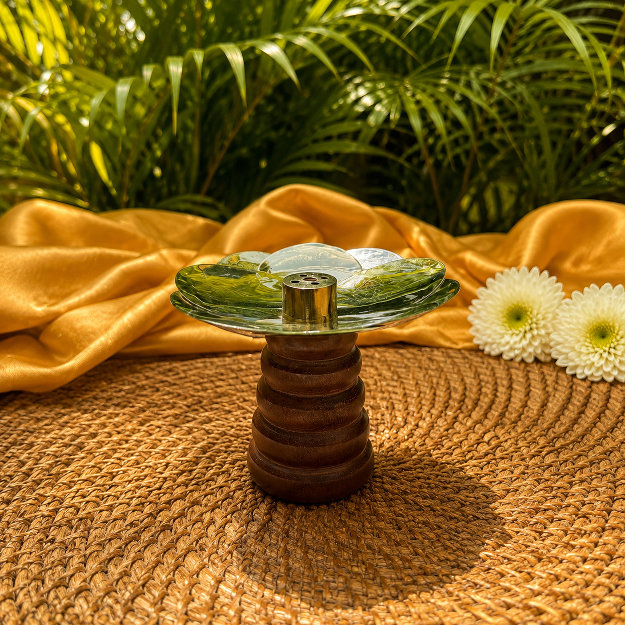 A glass and wood incense holder with a clear glass top and a ribbed brown wooden base, styled on a woven mat with yellow fabric and white flowers against a backdrop of green palm leaves.