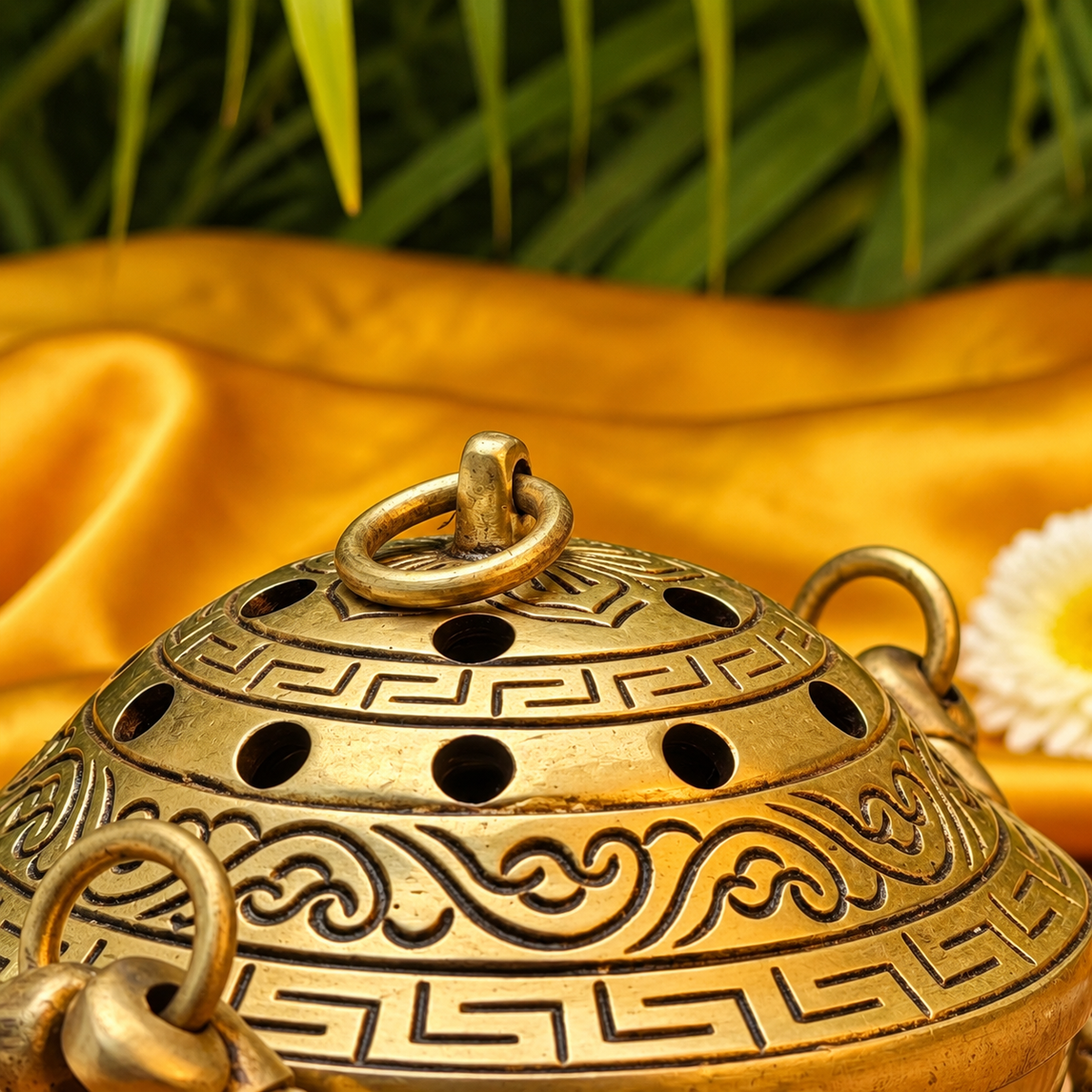 An ornate gold-toned metal incense burner with carved scrollwork and circular ventilation holes, shown close up on draped satin fabric with blurred green leaves in the background.