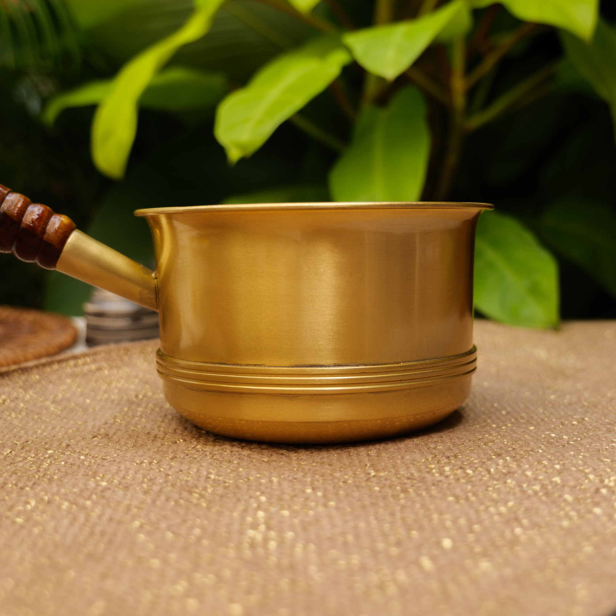 Bronze-colored pot with a wooden handle on a textured surface with green foliage in the background