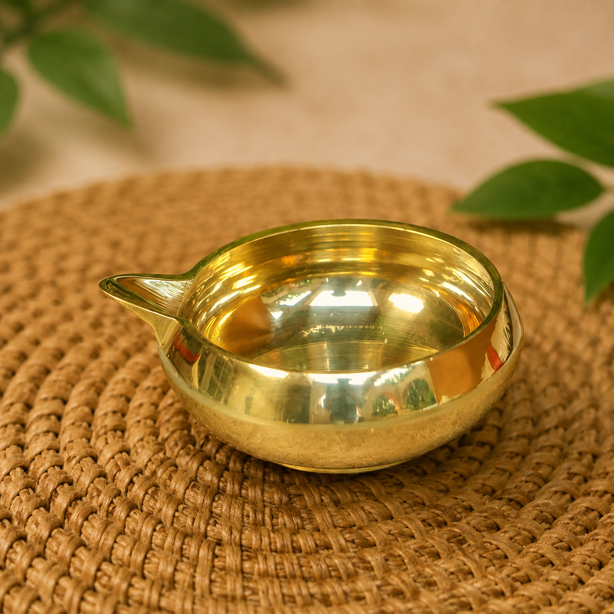 A small polished brass oil lamp with a spout, resting on a round woven straw mat with blurred green leaves in the background.