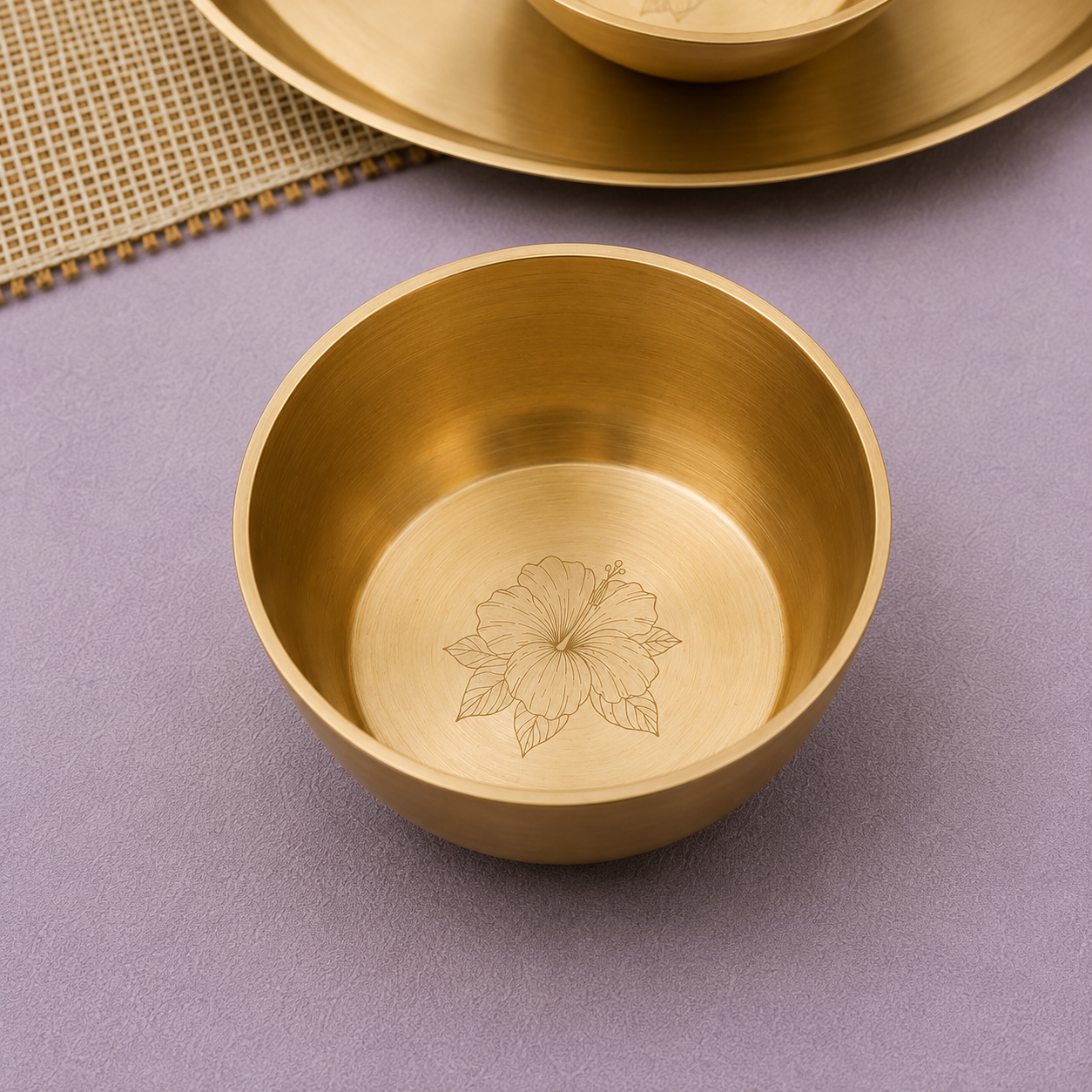 A brushed brass bowl with an engraved floral pattern inside, shown on a textured lavender surface with matching brass tableware partially visible in the background.