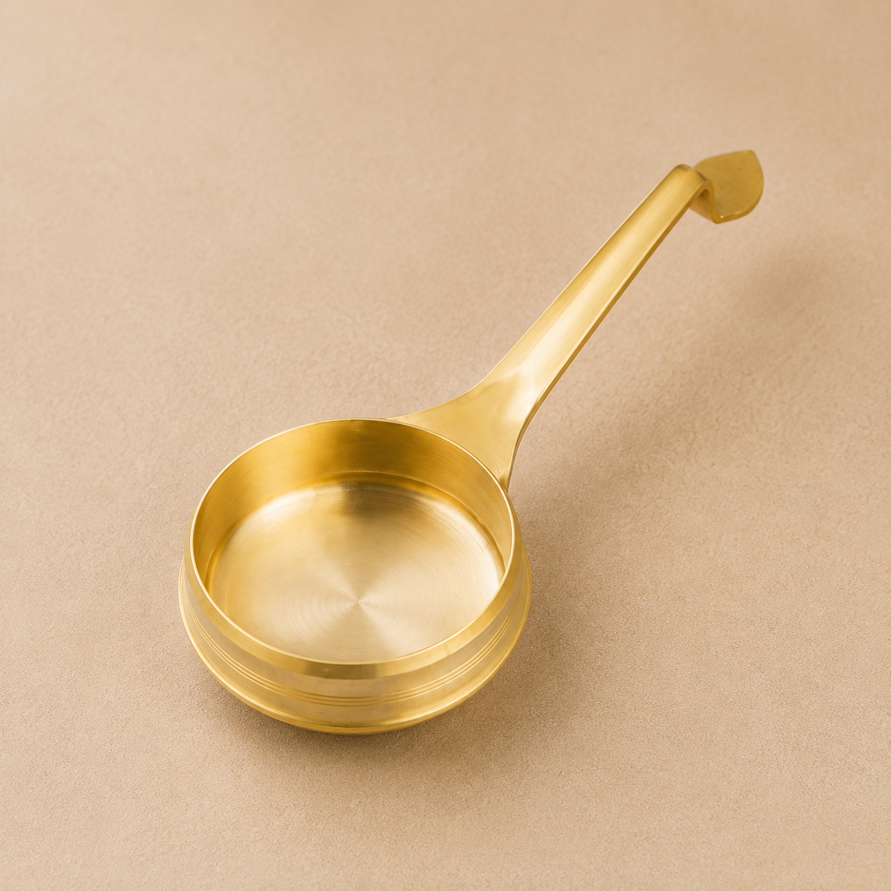 A polished brass ladle with a long handle, resting on a textured beige background.