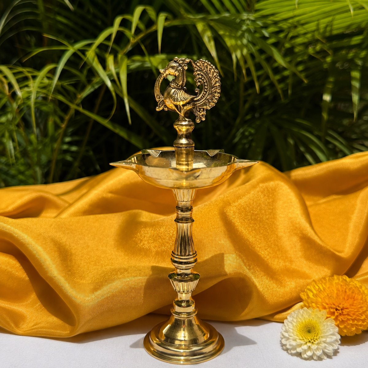 A brass oil lamp with a decorative peacock finial on top, styled on draped golden-yellow fabric with green palm leaves in the background and small flowers in the foreground.