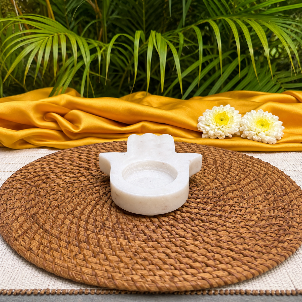 A small white stone hamsa hand holder sits on a round woven brown placemat, with a draped mustard-yellow fabric and green palm leaves in the background.