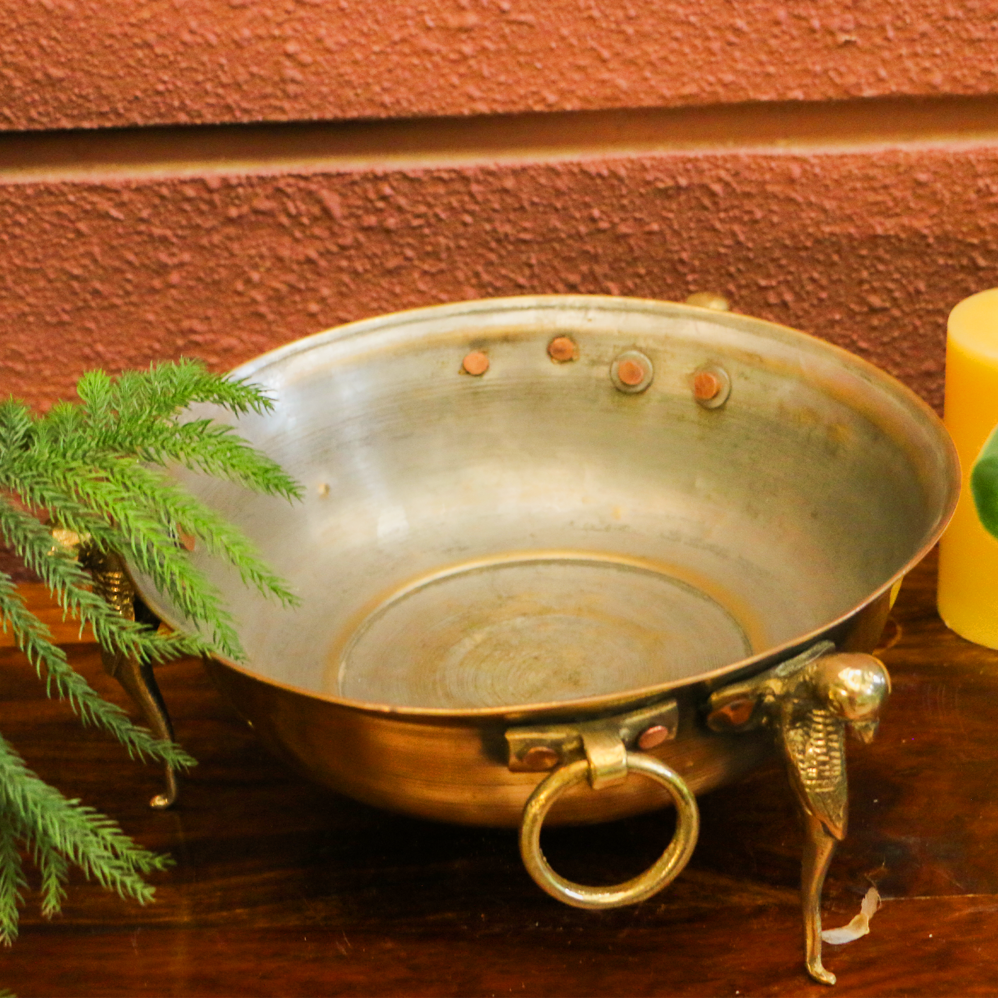 Bronze bowl with handles on a wooden surface with a plant and candle in the background