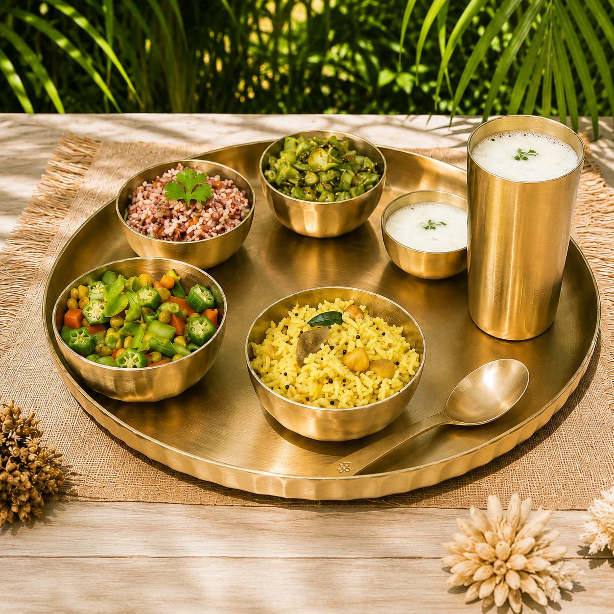 A brass serving tray set on a wooden table with a woven placemat, holding five bowls of food and a tall cup of milk, with a spoon on the tray. The background shows blurred green foliage.