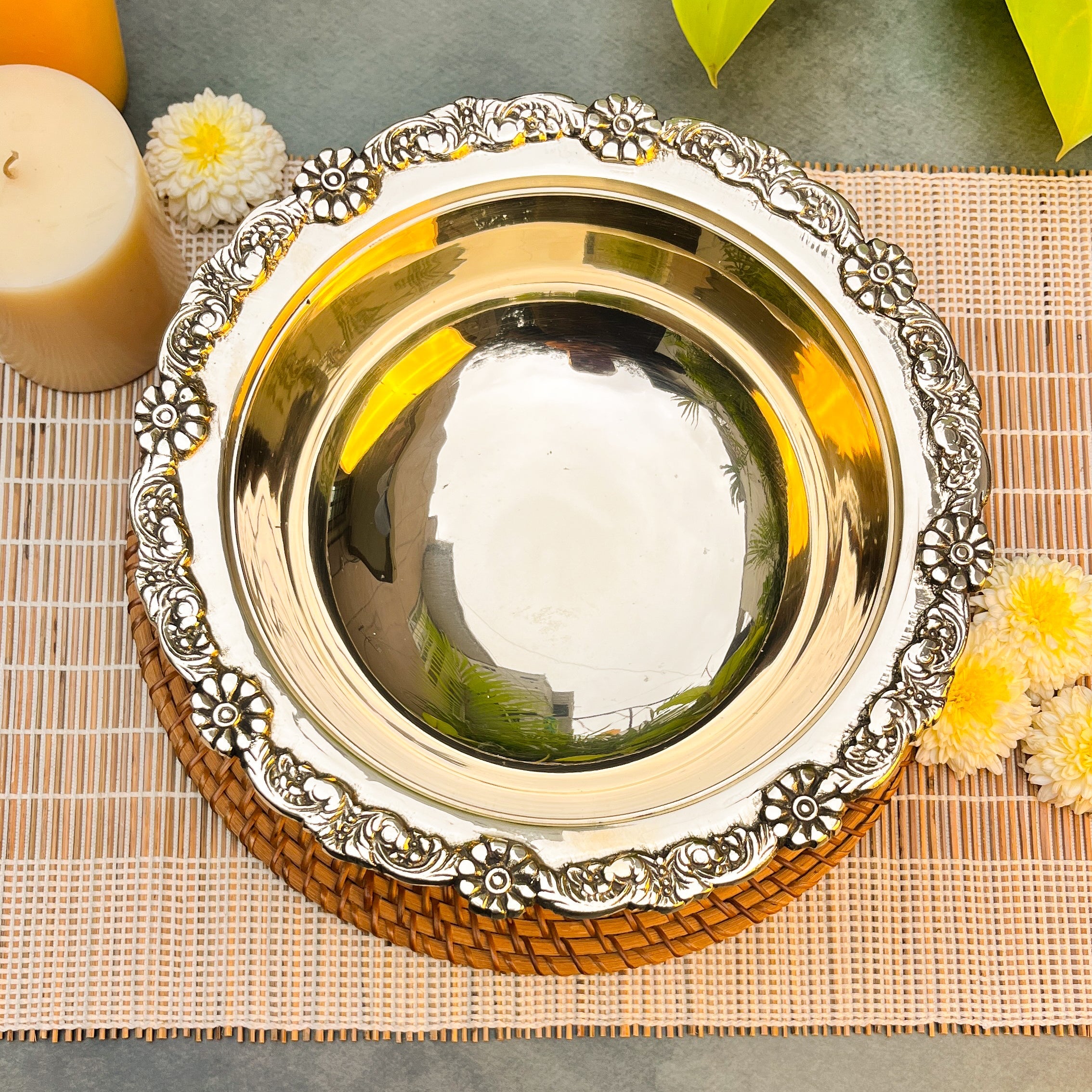 Decorative silver and gold plate on a woven mat with flowers and a candle in the background