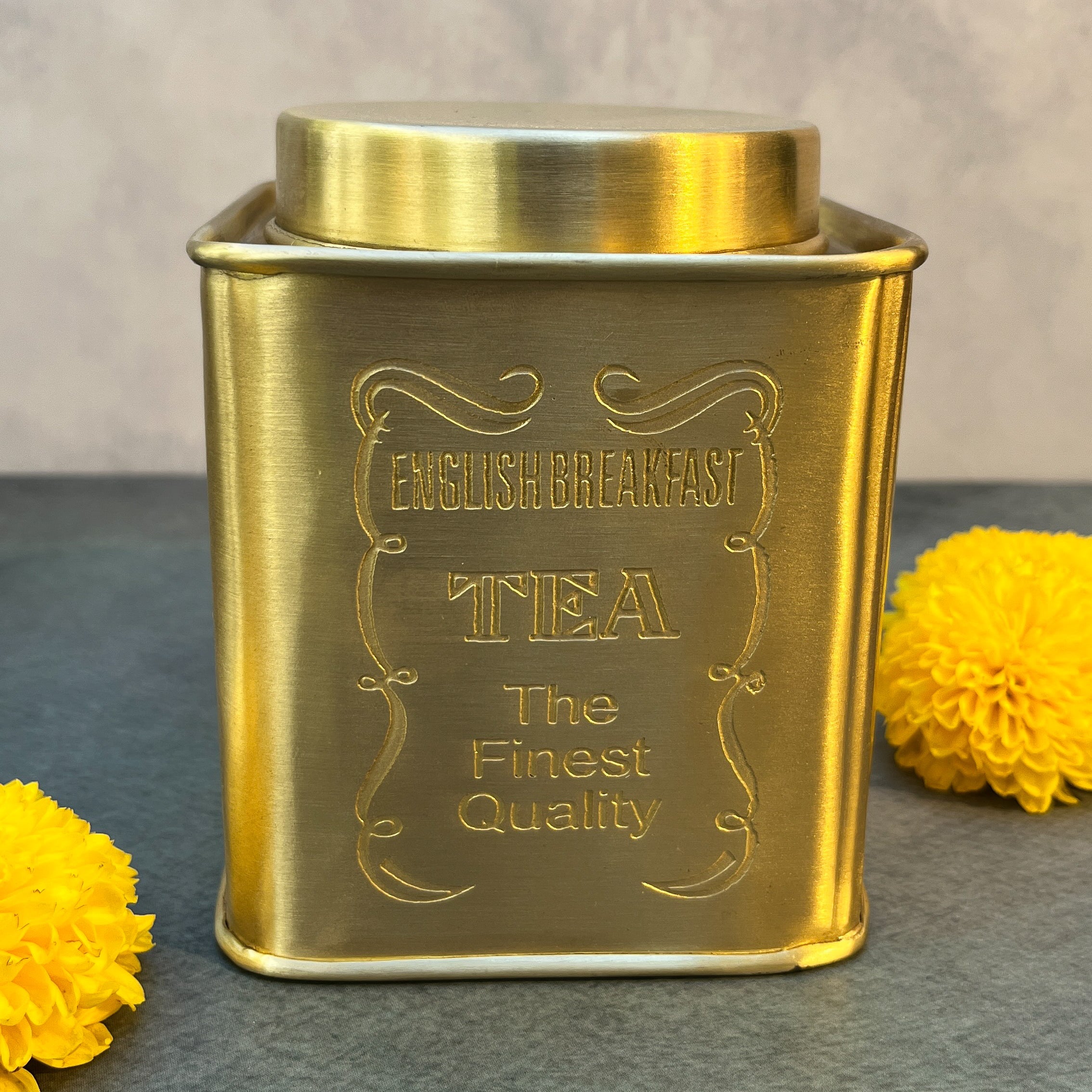 Gold tea canister with 'English Breakfast Tea' text on a gray surface with yellow flowers.
