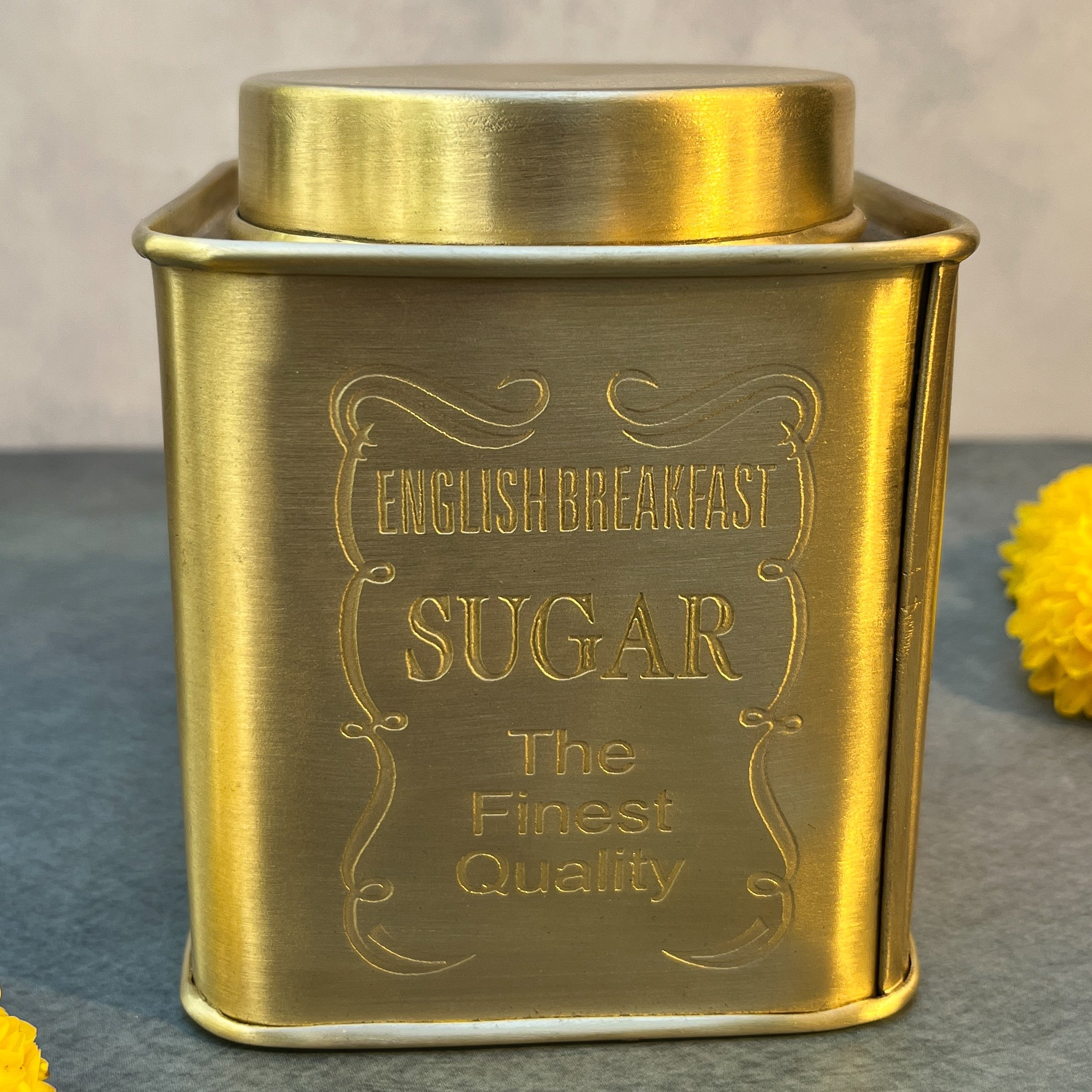 Gold-colored container labeled 'English Breakfast Sugar The Finest Quality' on a gray surface with a yellow flower.