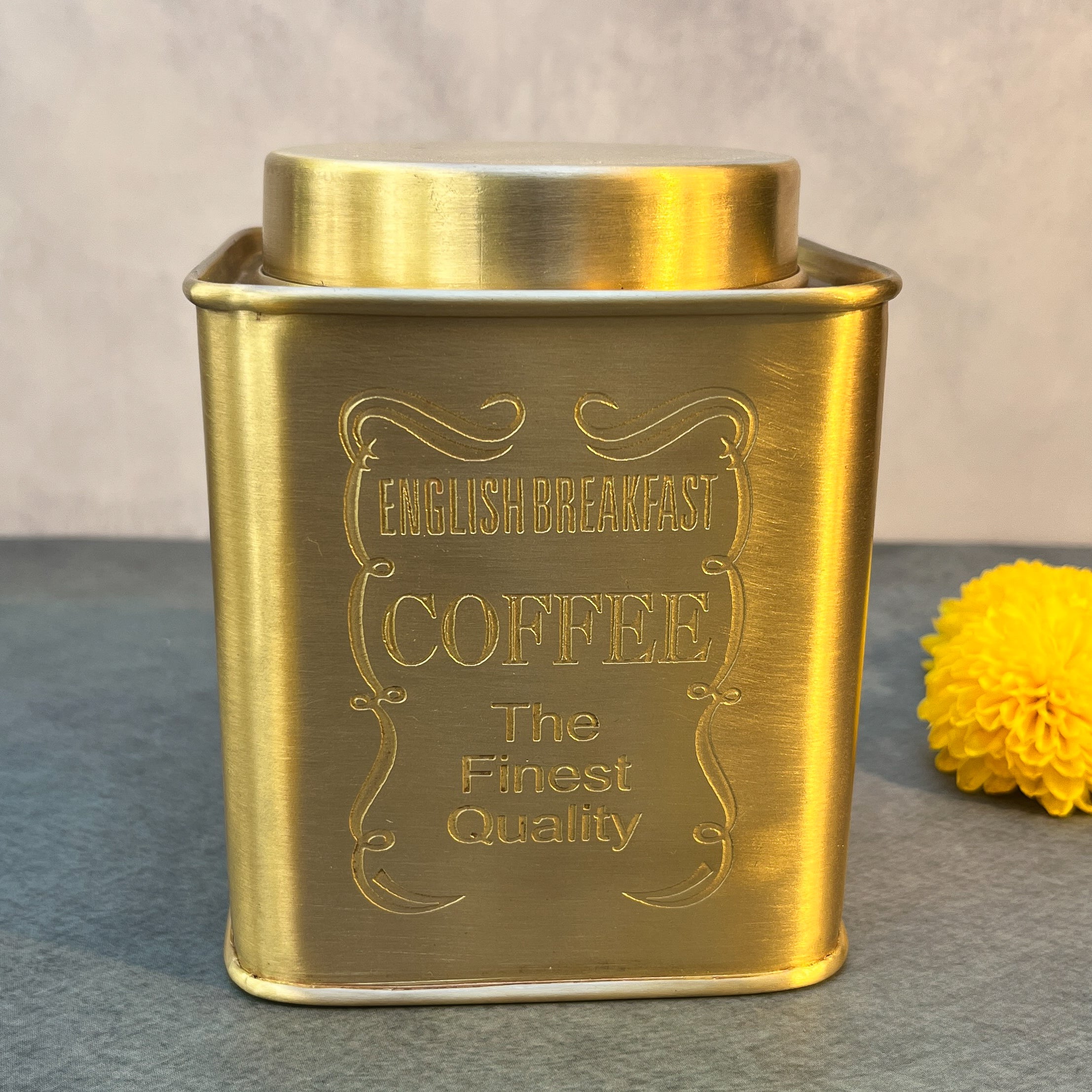 Gold coffee canister with 'English Breakfast Coffee' text on a gray surface with a yellow flower.