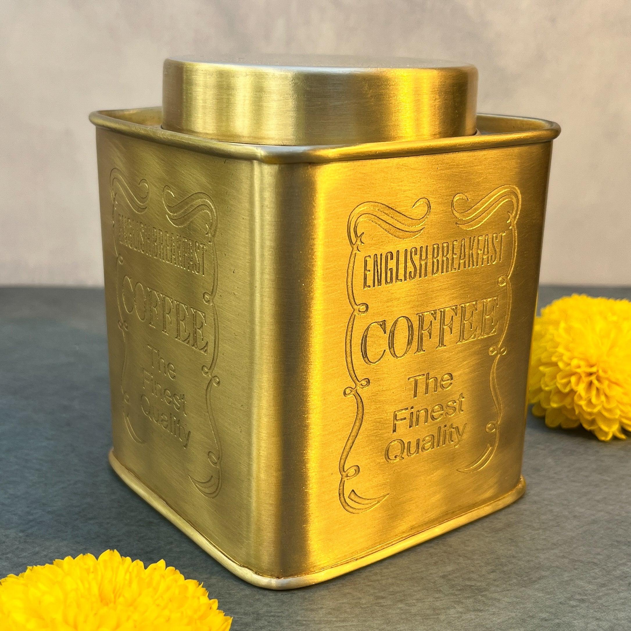 Gold coffee canister with branding text on a gray surface with yellow flowers.