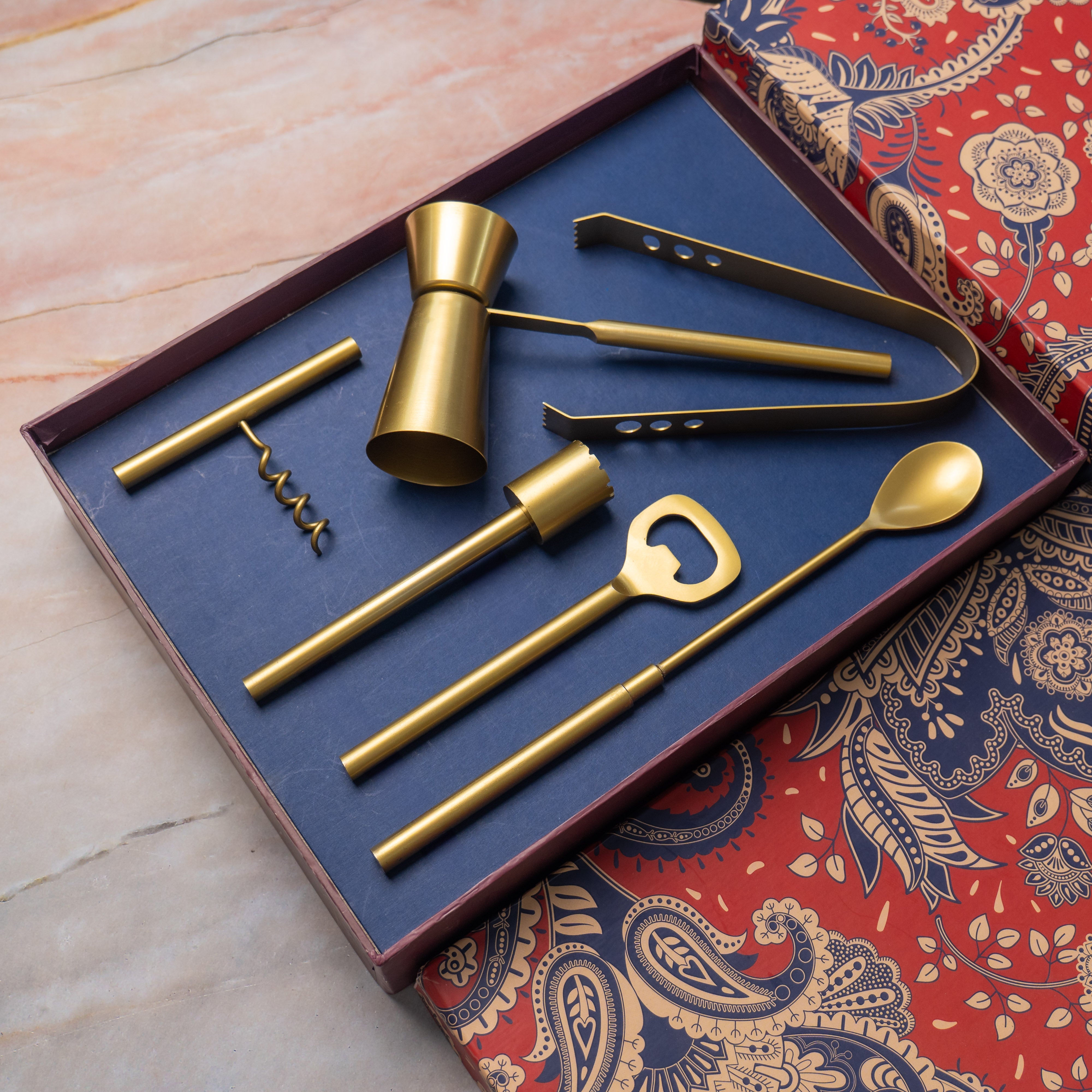 Set of gold bar tools including a corkscrew, jigger, and bottle opener on a blue tray with a decorative box in the background.