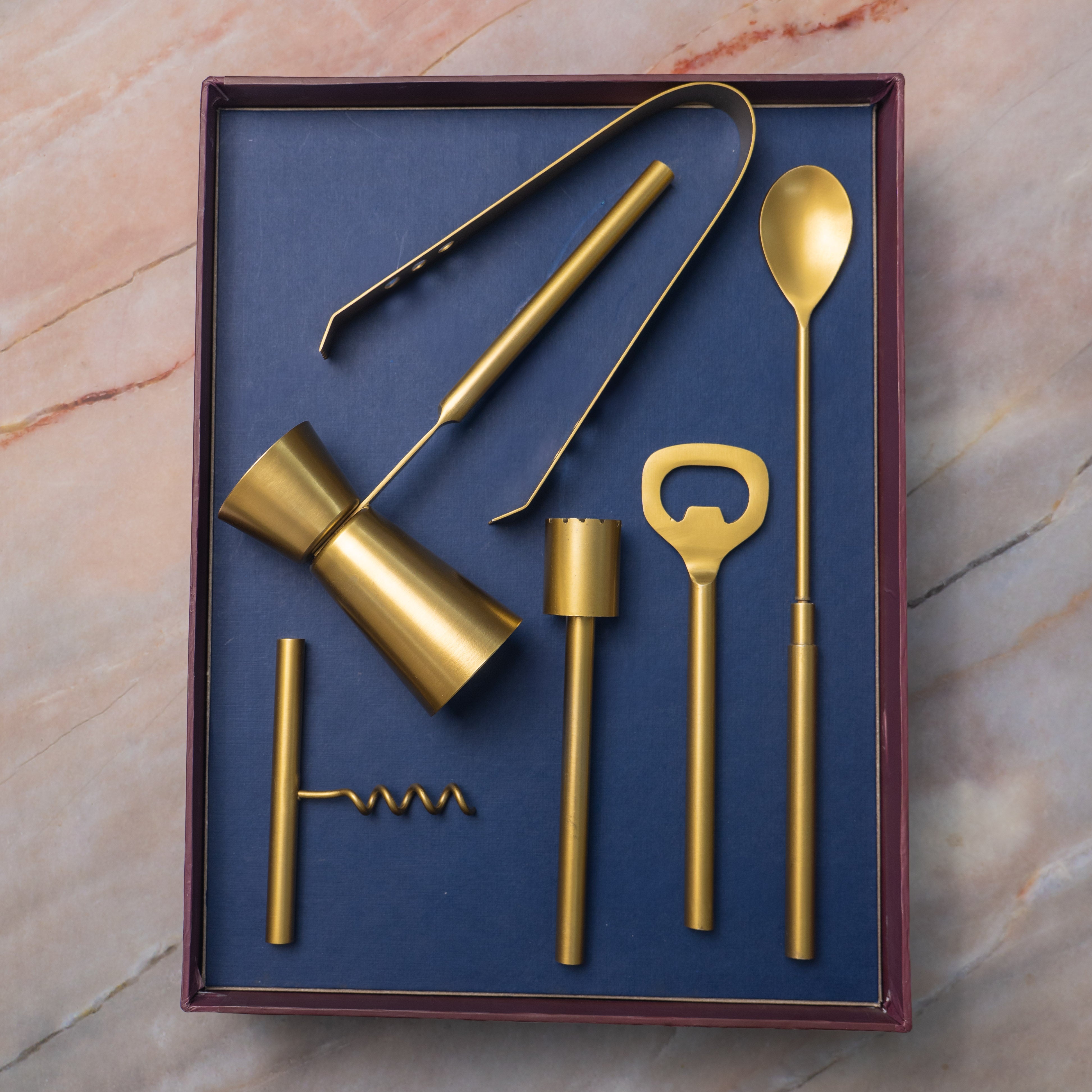 Set of gold bar tools including a jigger, spoon, and bottle opener in a box on a marble surface.
