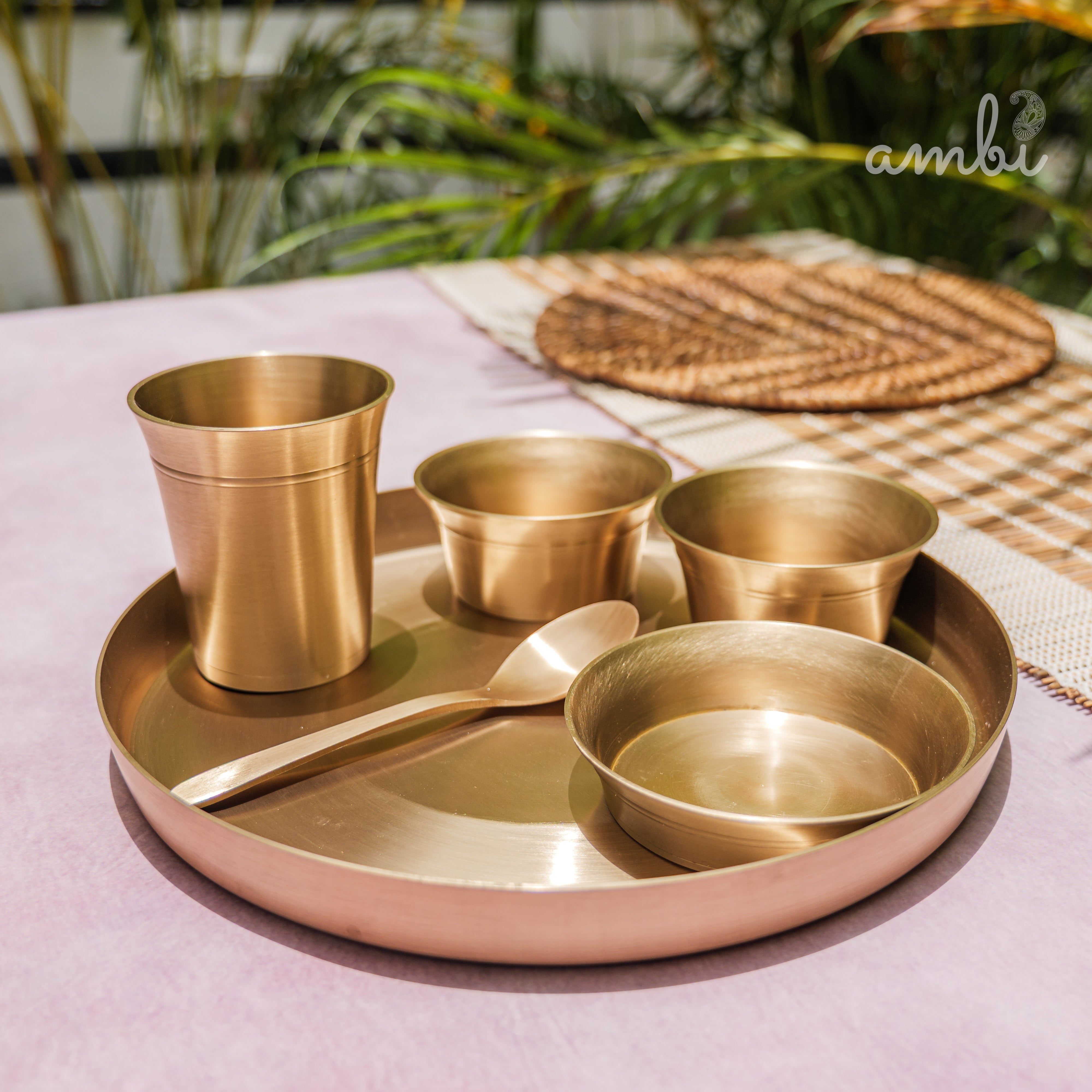 Set of gold-colored dining utensils including a tray, cups, bowls, and a spoon on a pink surface with plants in the background.