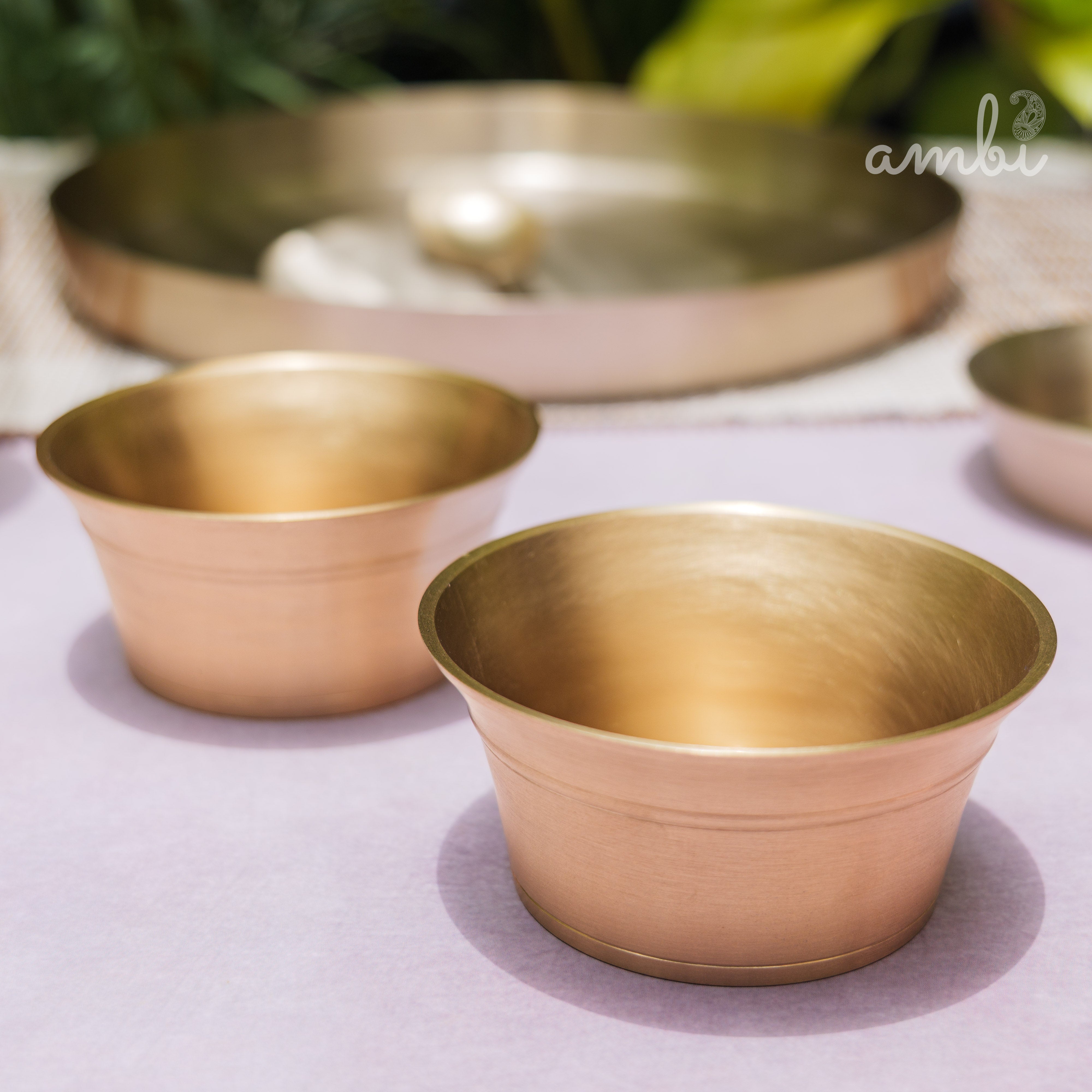Two brass bowls on a light purple surface with 'ambi' branding in the corner.