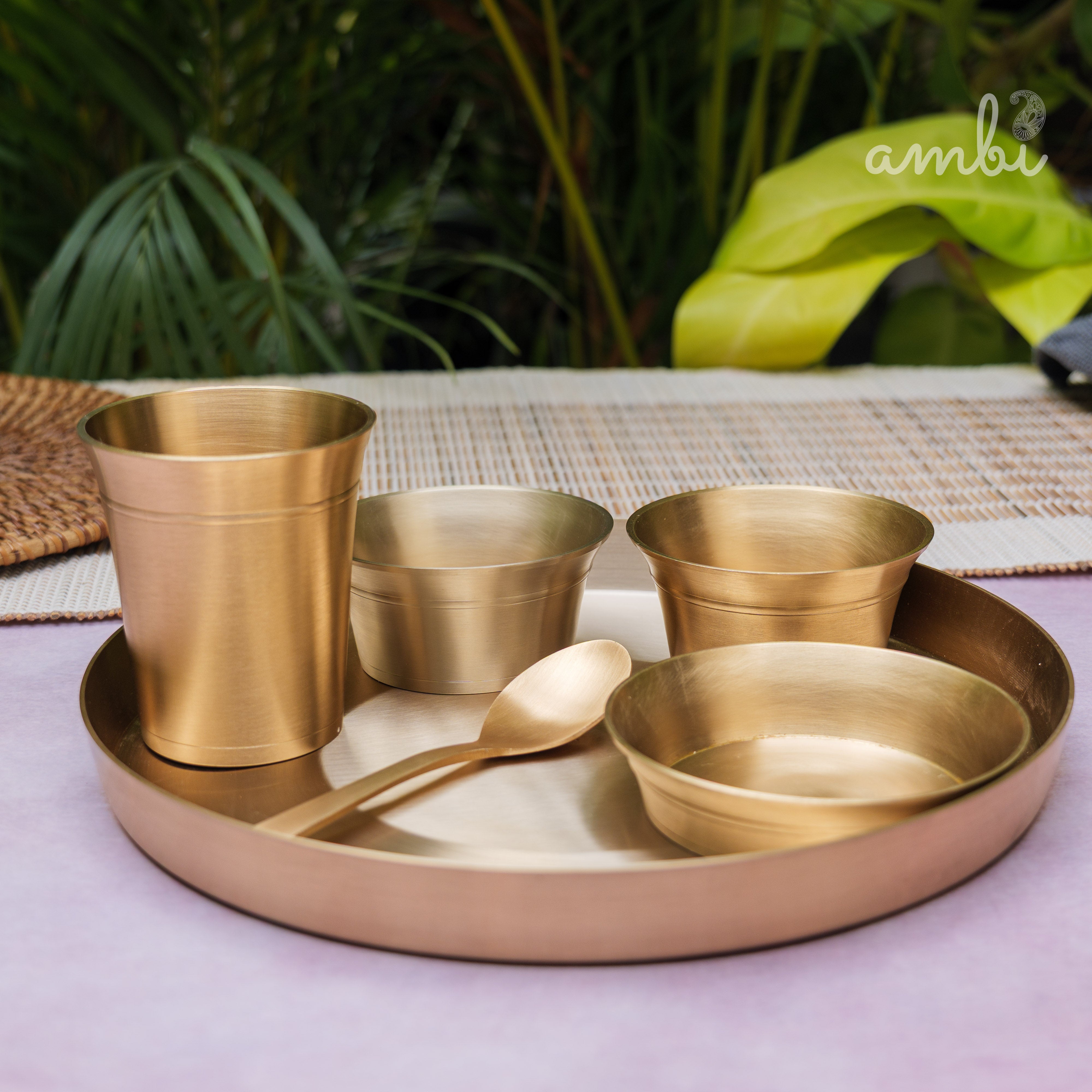 Set of gold-colored dining utensils including cups, bowls, and a spoon on a tray with a green leafy background.