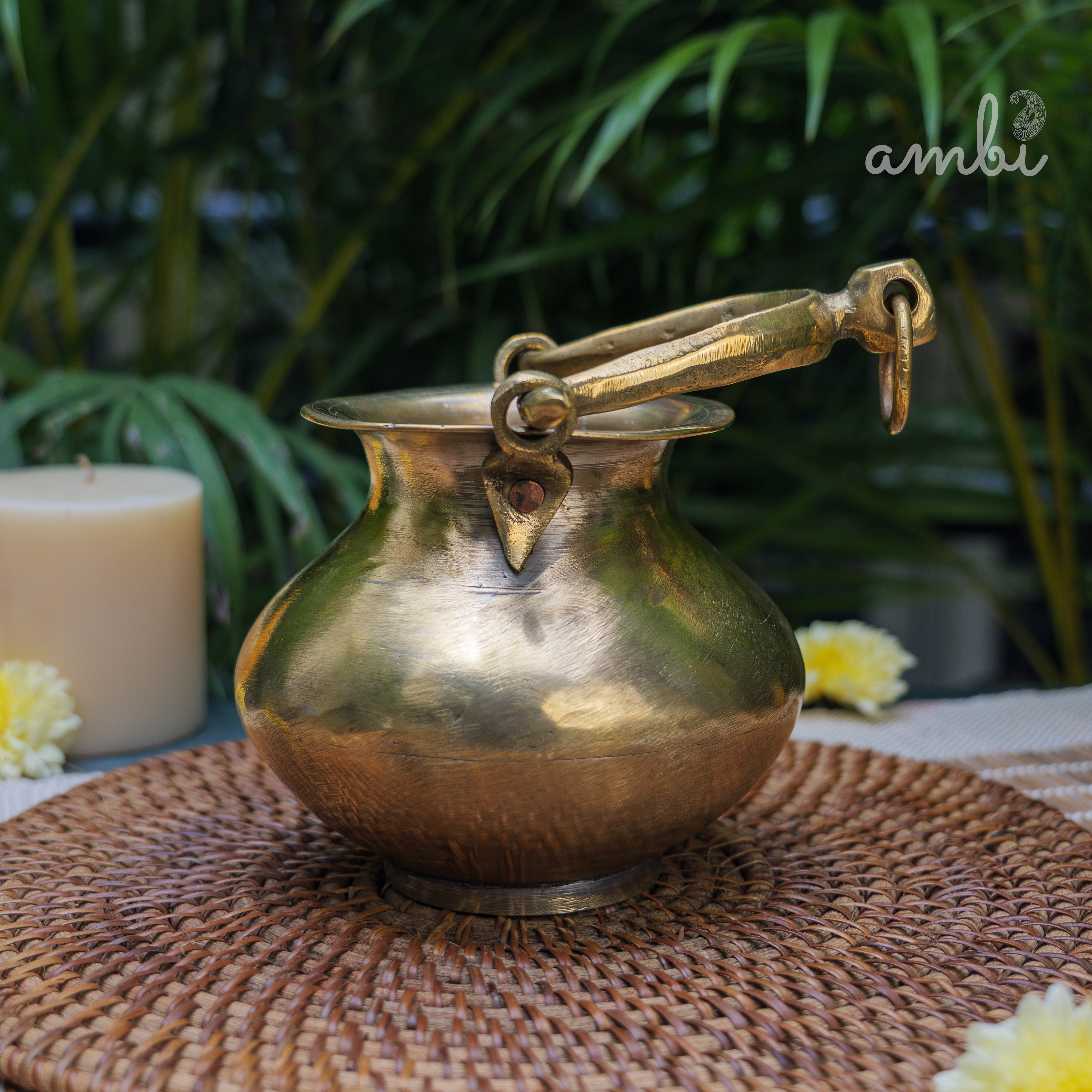 Brass pot with handle on a woven mat with plants and candle in the background