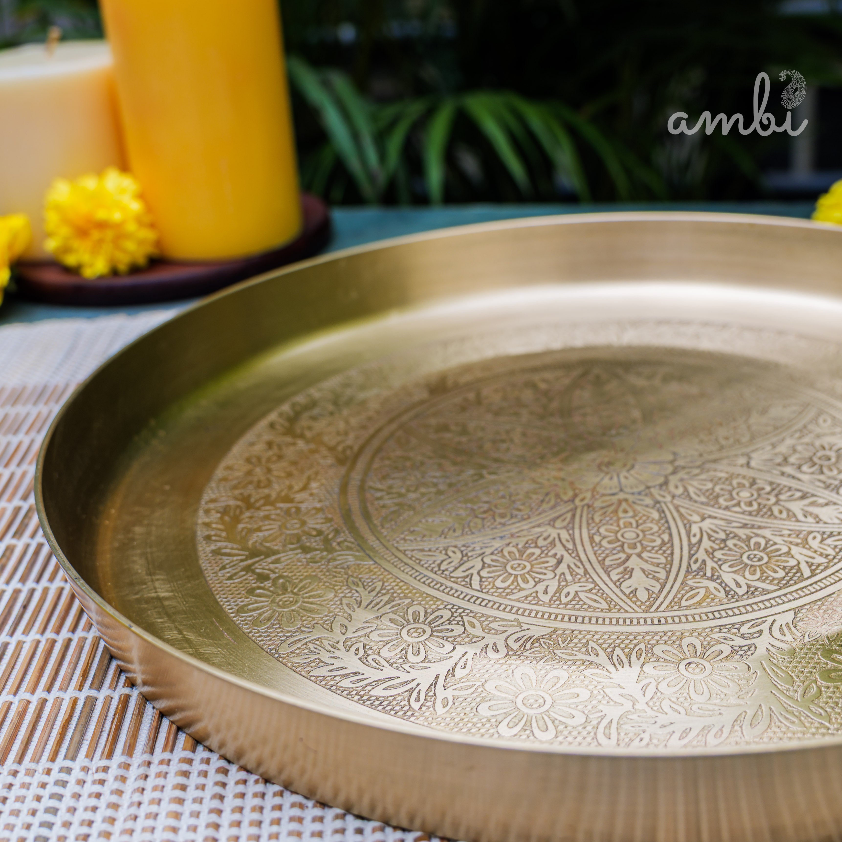 Decorative brass tray with intricate patterns on a table with flowers and a candle in the background.