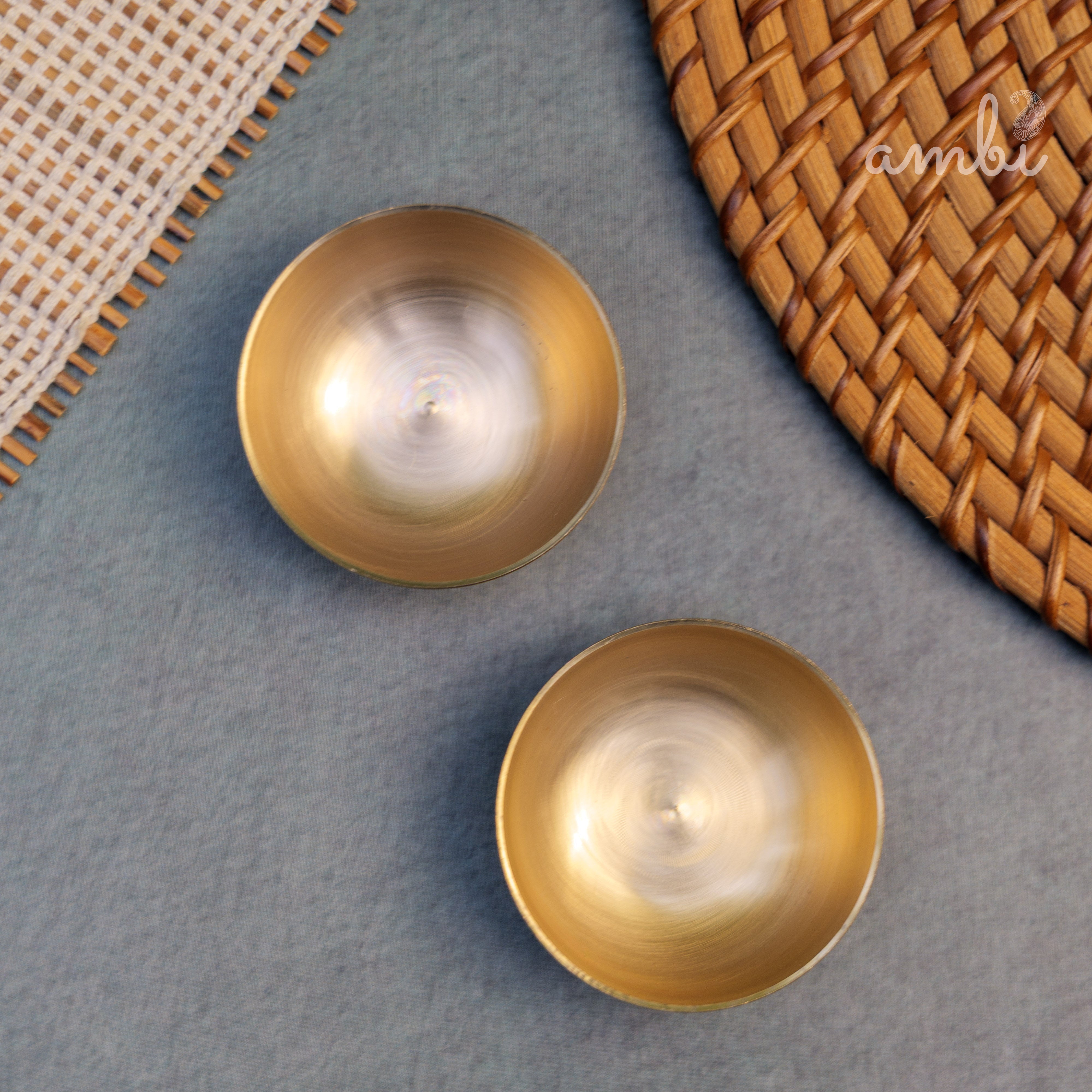 Two gold candles on a gray surface with a woven mat in the background, featuring the 'ambi' brand.