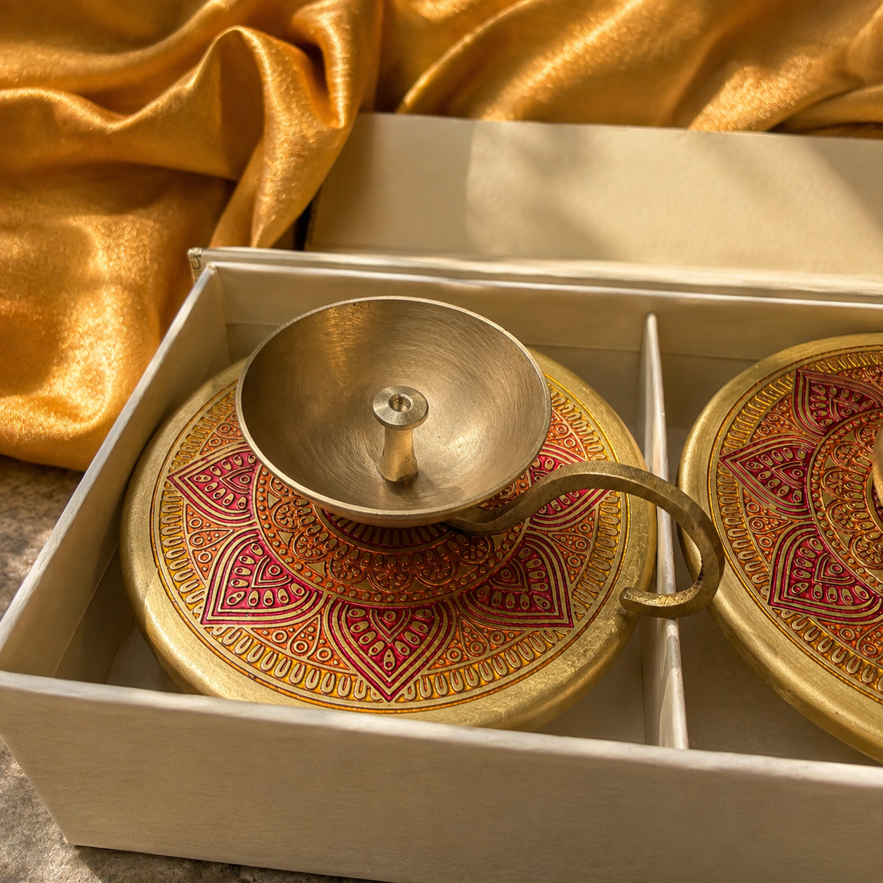 A brass-toned metal cup with a matching saucer lid decorated with red and gold mandala patterns, presented inside a cream-colored gift box with draped gold fabric in the background.