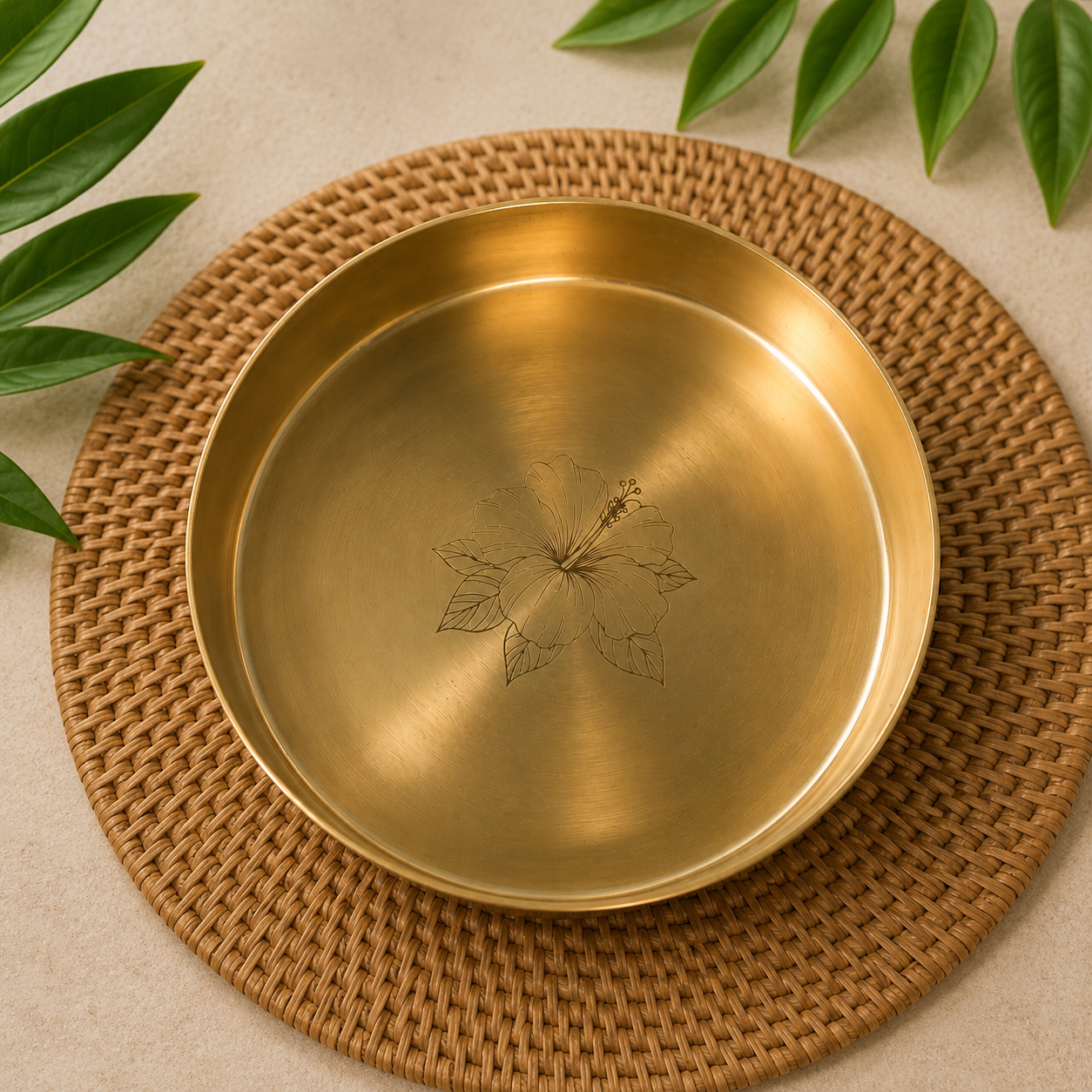 A round brushed brass plate with an engraved hibiscus flower design in the center, resting on a woven brown placemat with green leaves in the corners.