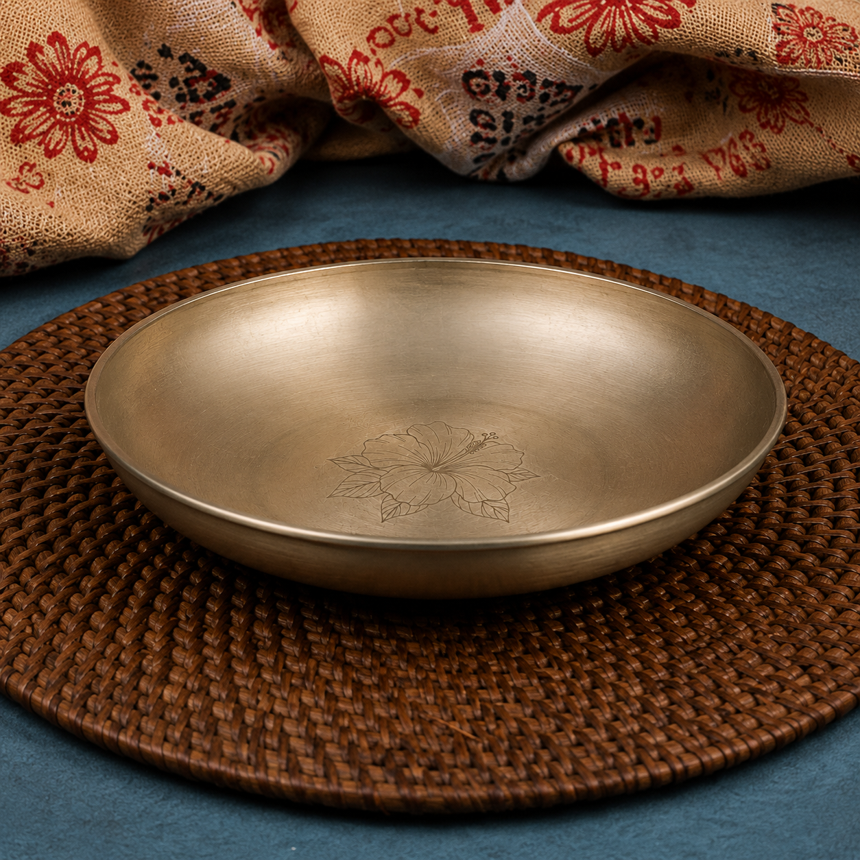 A round metallic bowl with a brushed silver finish and an engraved floral motif, displayed on a brown woven placemat with a patterned fabric backdrop.