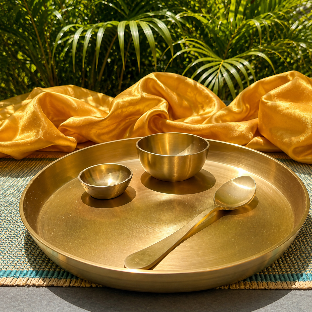 A brass tray with a matching spoon and two small bowls, styled on a woven mat with draped gold fabric and green palm leaves in the background.