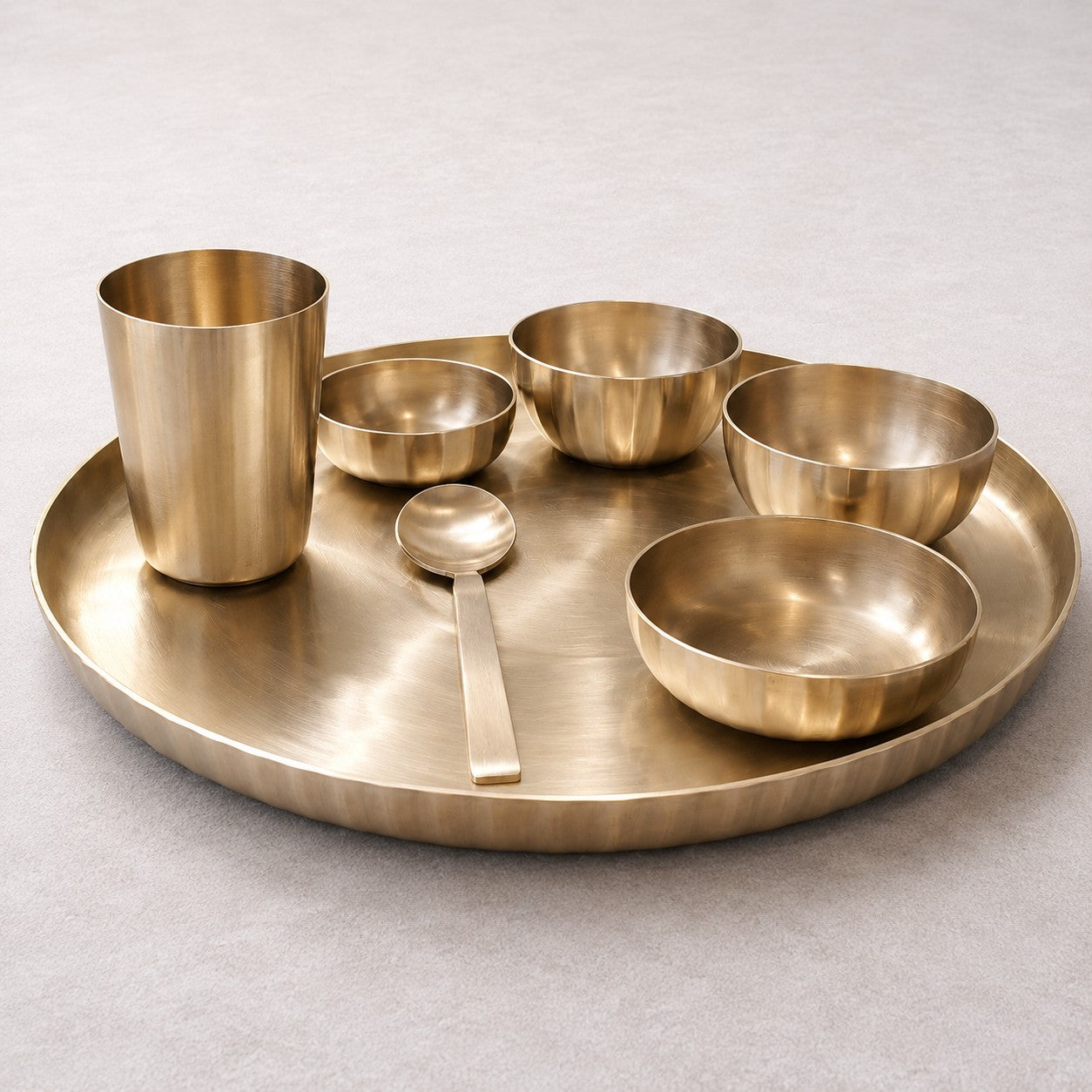 A brass-colored metal serving set including a large round tray, a tumbler, a spoon, and four bowls, arranged on a neutral light gray surface.