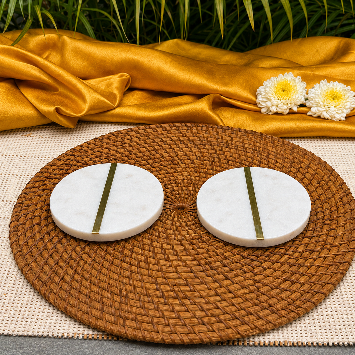 Two round white marble coasters with vertical gold metal accents, arranged on a brown woven placemat with a draped gold fabric and white flowers in the background.